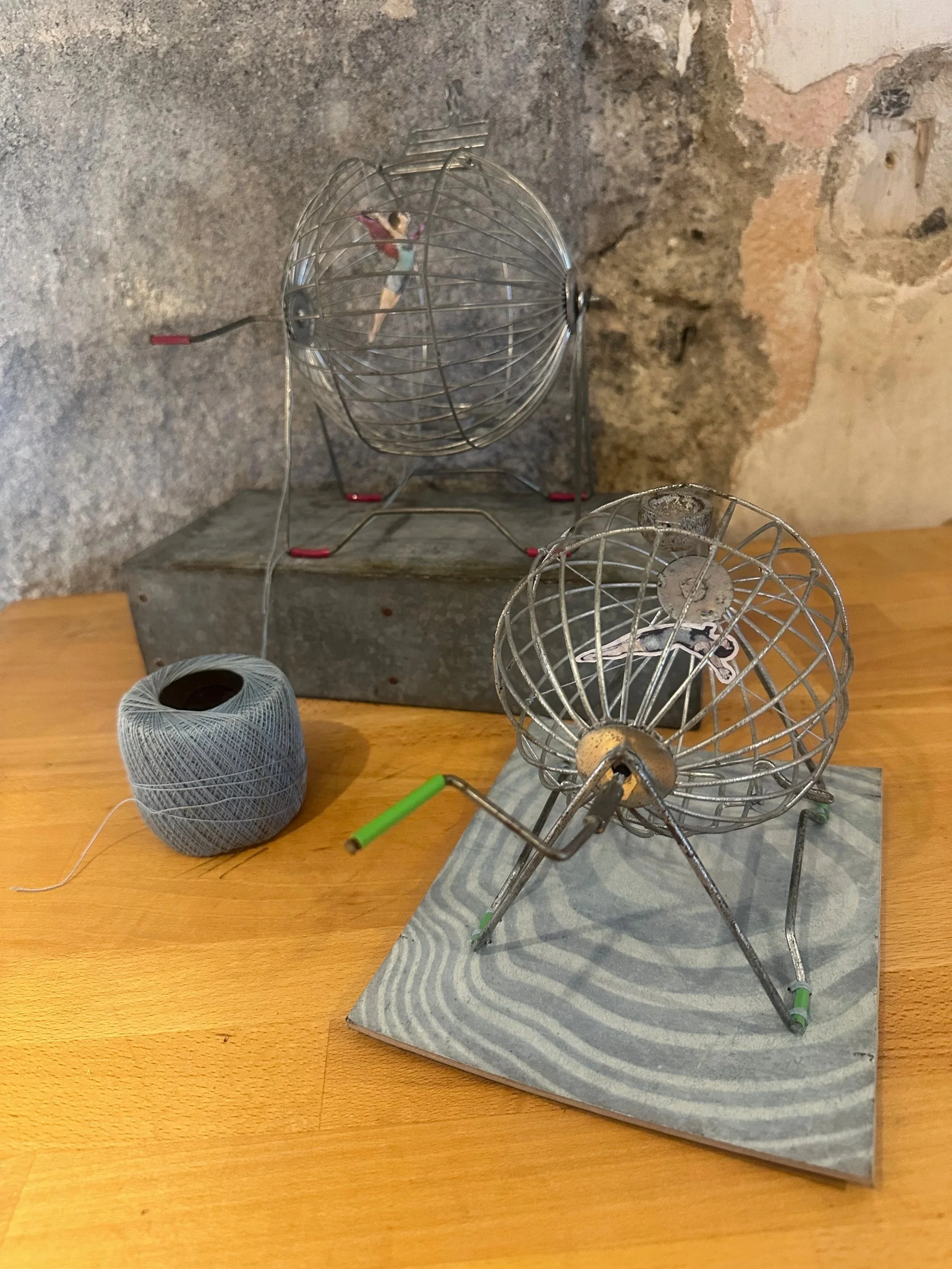A metal hamster wheel with a small figurine inside, set on a wooden surface with a ball of gray thread nearby and a textured gray background.