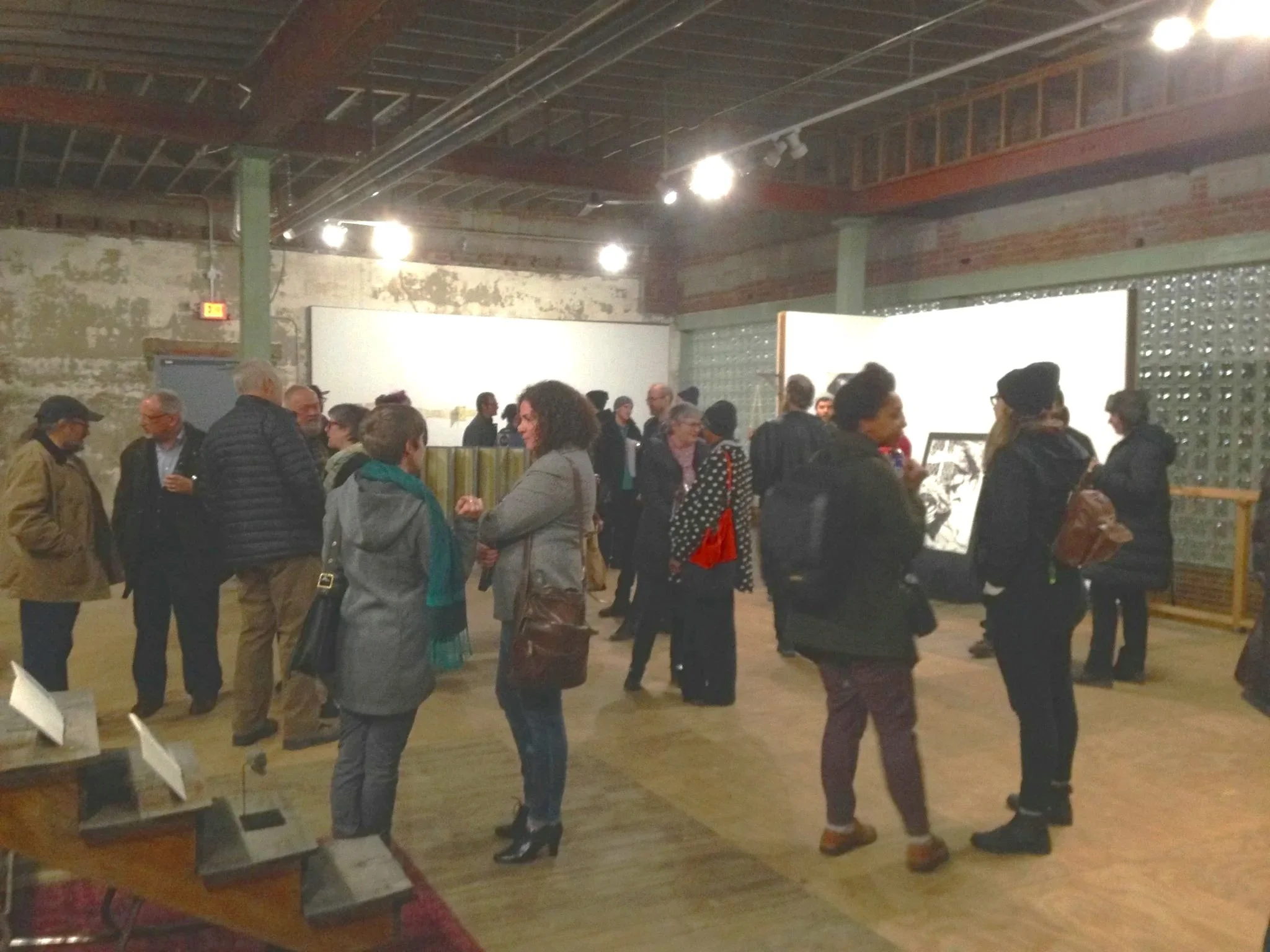 A group of people gathered in an indoor art gallery,  studio space, Holding House, 3546 Detroit Michigan Ave, looking at artwork displayed on the walls.