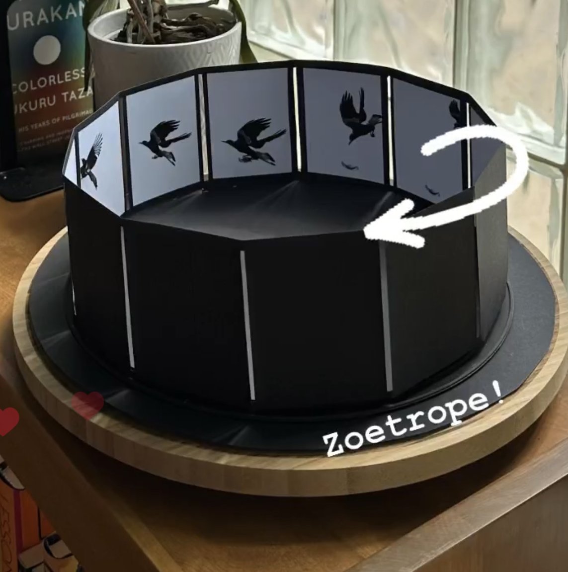 A circular, black photo display stand with silhouette images of flying birds on its panels. The stand is on a wooden surface near a window, with a plant and a cleaning product in the background. The word "Zoetrope!" is written on the stand.