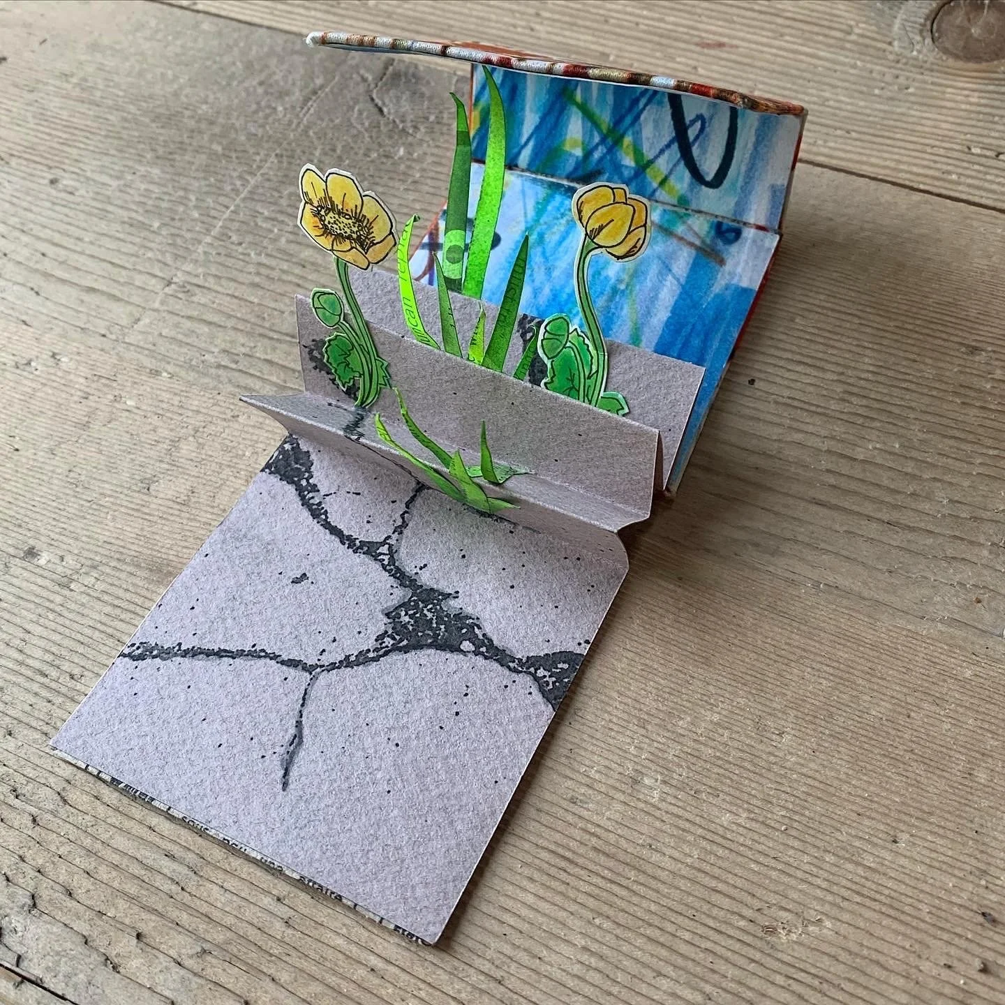 A handmade 3D paper craft of yellow flowers with green stems and leaves, set against a reflective mirror paper background, on a wooden surface.