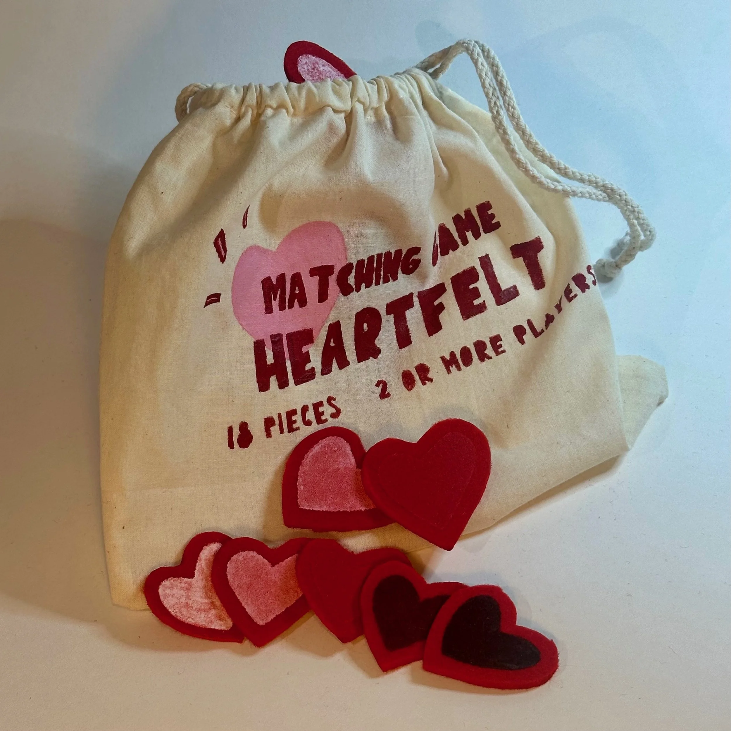 A beige drawstring bag with red text and pink hearts, containing felt heart shapes in various shades of red and pink, with some hearts spilling out of the bag.