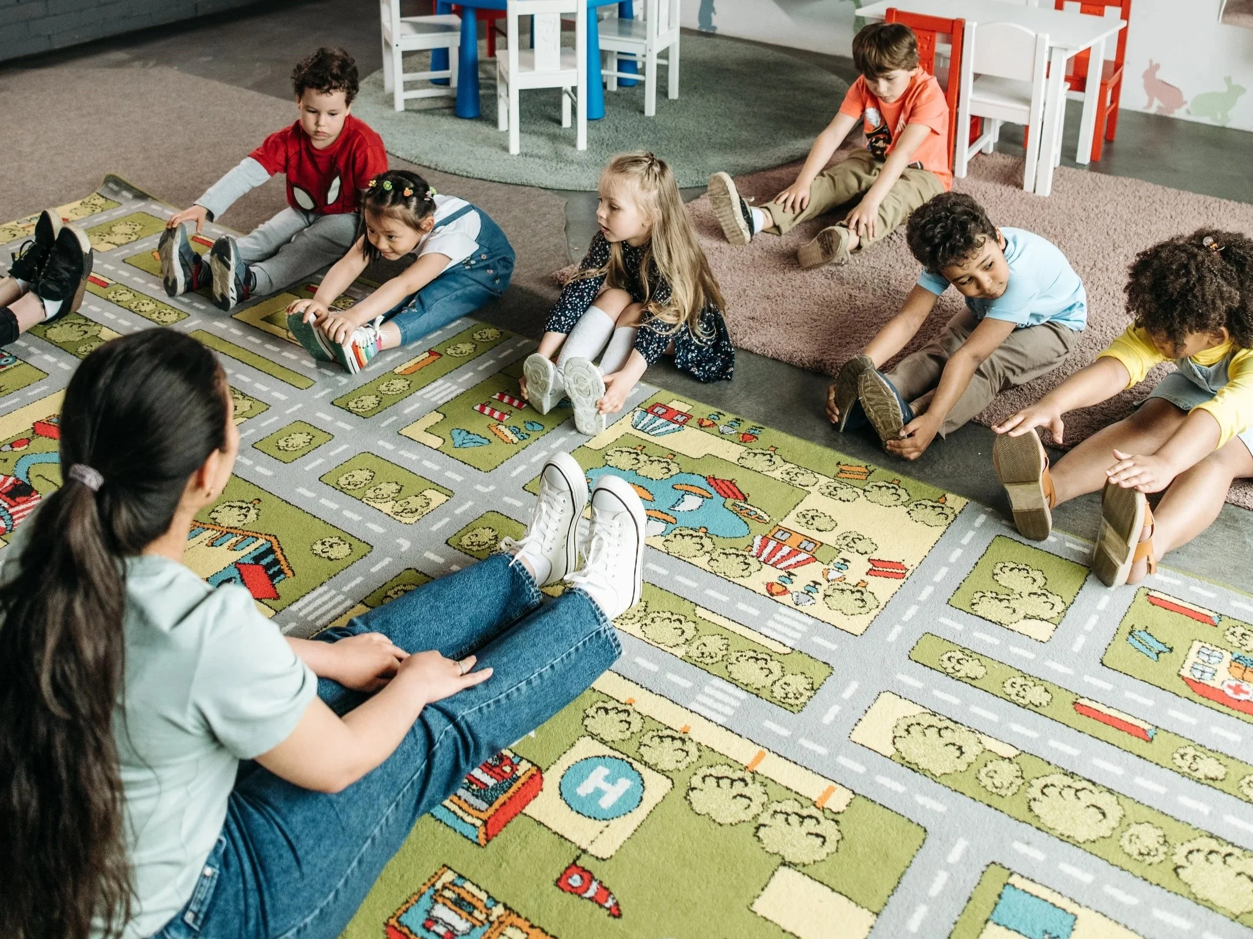 Teacher in classroom on meeting rug teaching a group of kids to touch their toes.