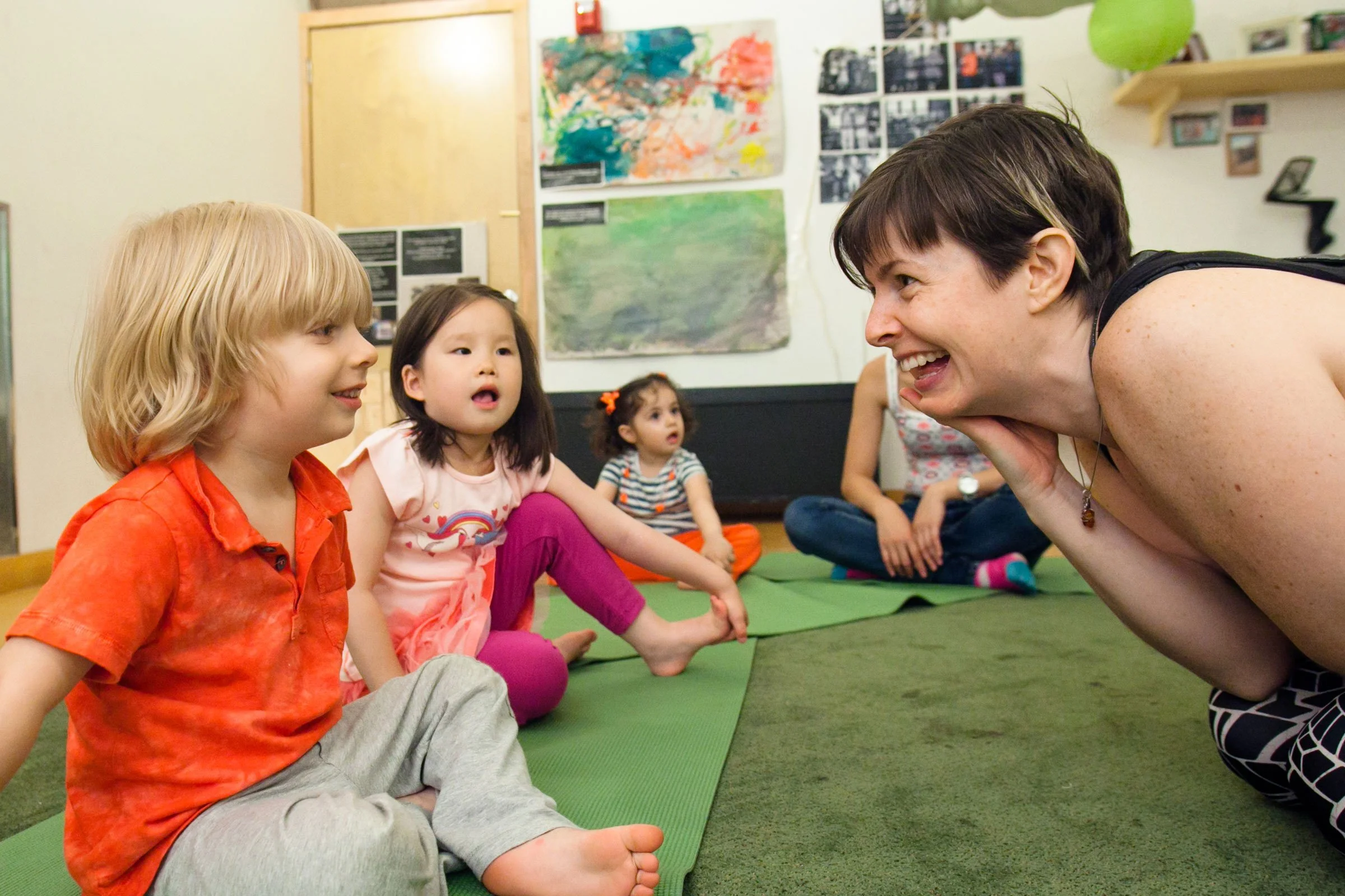 Young girl and adult woman blowing on a small white ball as part of a playful breathing exercise in a kids yoga and mindfulness session, surrounded by wooden blocks in a classroom setting.