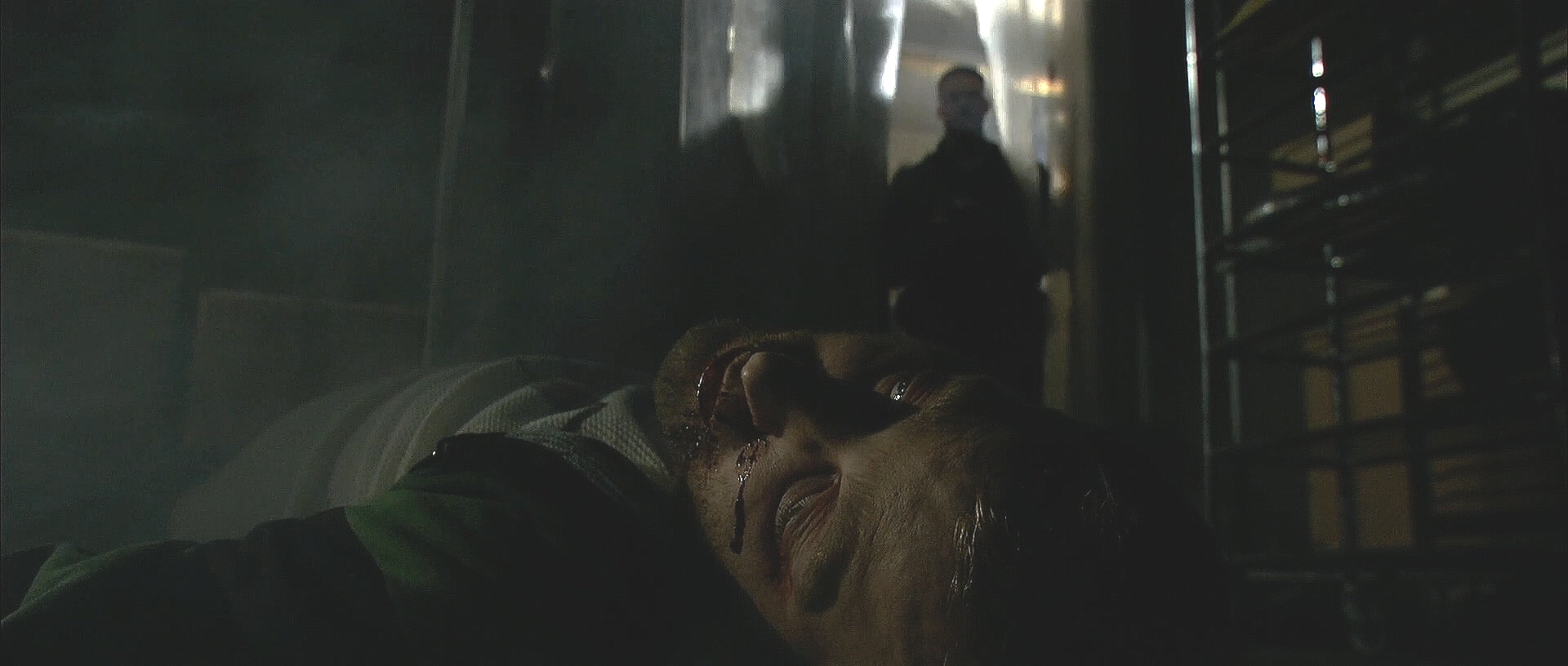 A person lying on the ground with blood on their face, in a dark, industrial setting. Two figures stand in the background, partially silhouette, looking towards the person on the ground.