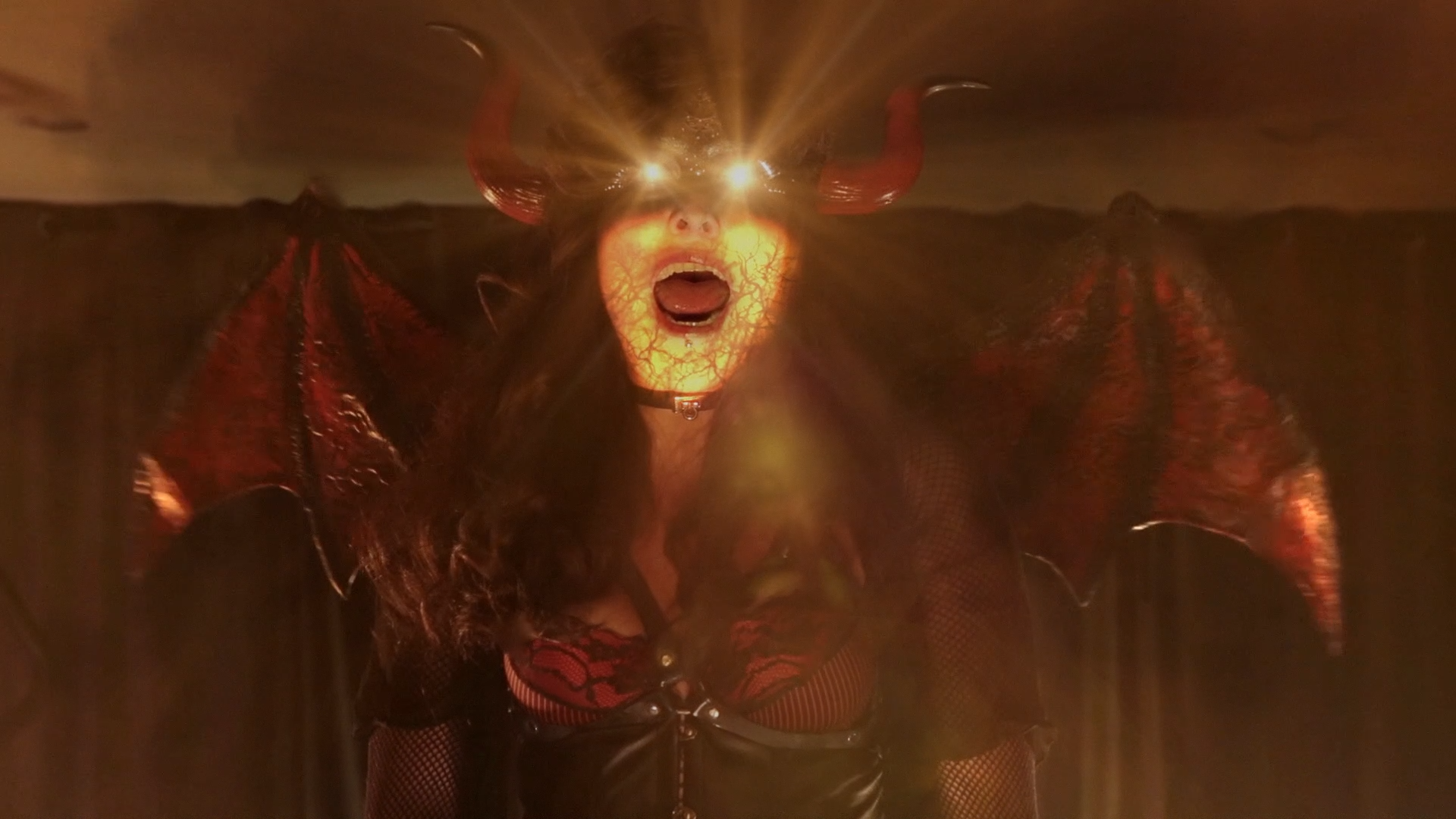 A person dressed in a devil costume with horns, wearing a cloak, with bright lights behind their head, mouth open, and looking at the camera.