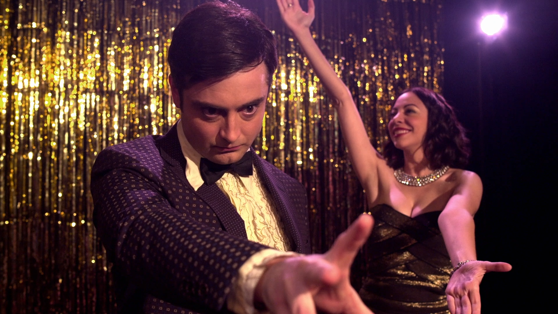 A man in a tuxedo with a bowtie and a woman in a strapless dress with jewelry dancing at a party with a gold sequin backdrop.