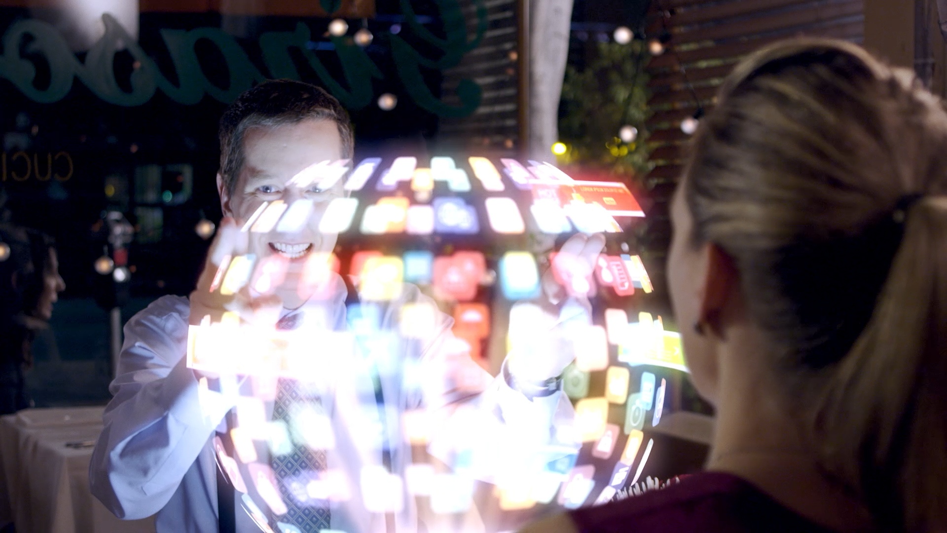 A man in a white shirt and patterned tie smiling and holding a digital device, with holographic app icons surrounding him, while a woman with tied-back hair looks on in a dimly lit outdoor setting at night.
