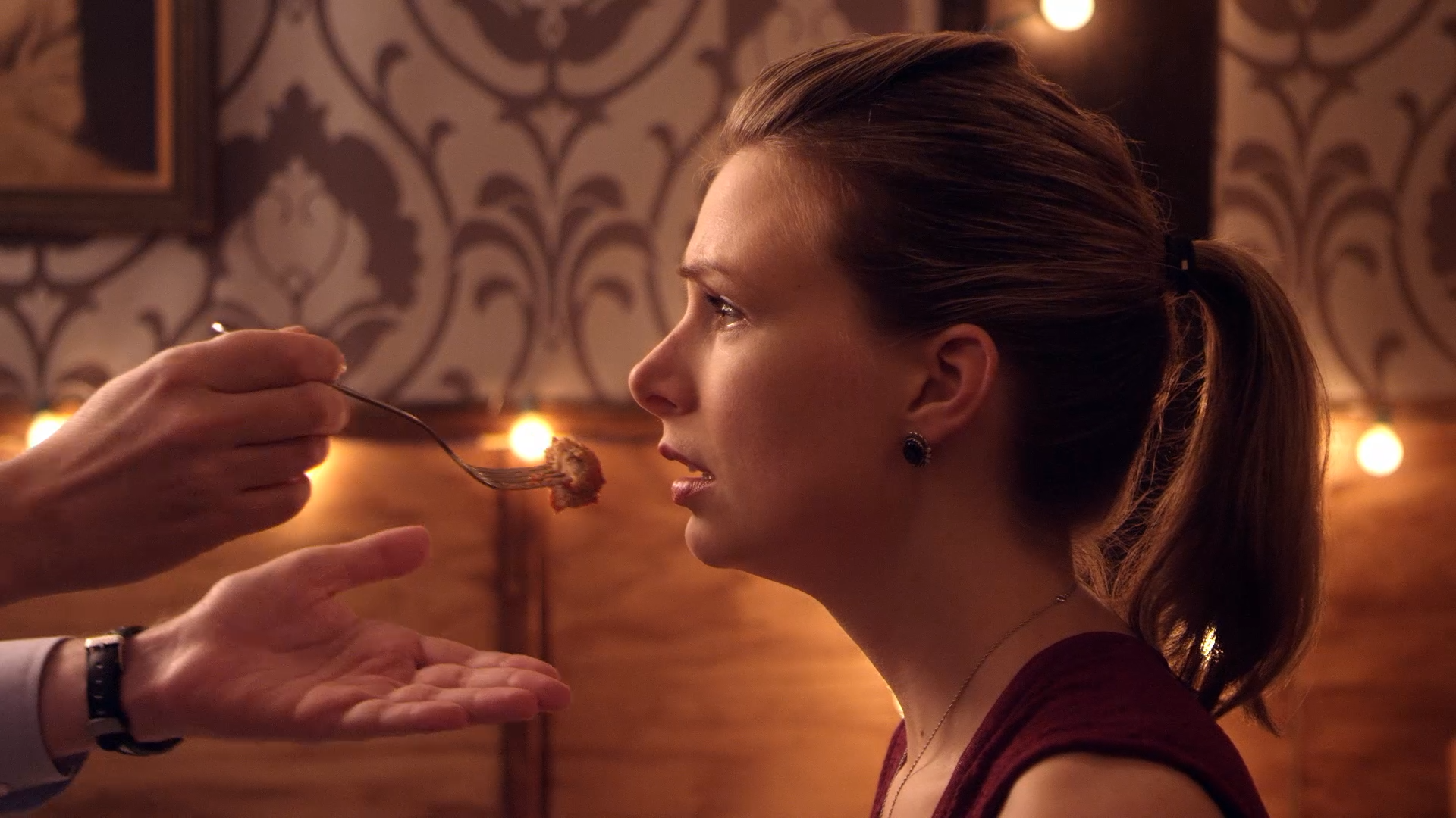 A woman with a ponytail receiving a forkful of food in a dimly lit restaurant with decorative wallpaper and warm lights.