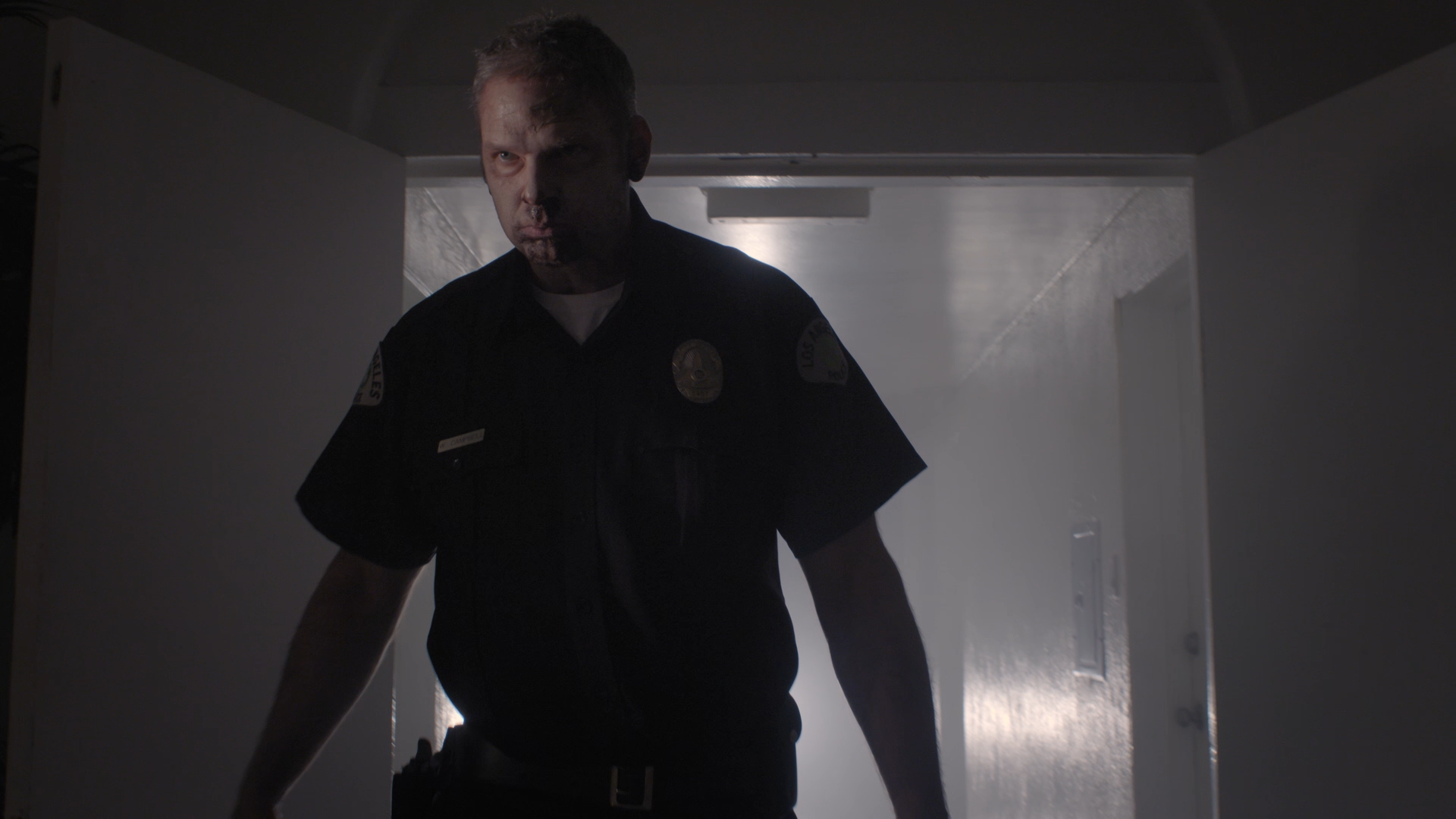 A police officer walking through a dimly lit hallway, dressed in a dark uniform with visible badges, with a serious expression and shadows cast on his face.