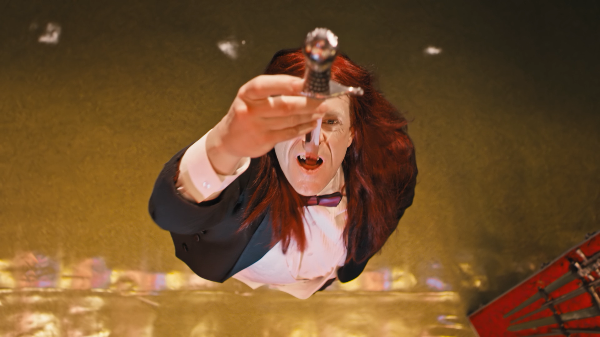 A man with long red hair and a tuxedo, with an open mouth, holding a sword pointing downward, seen from below, against a blurred golden background.