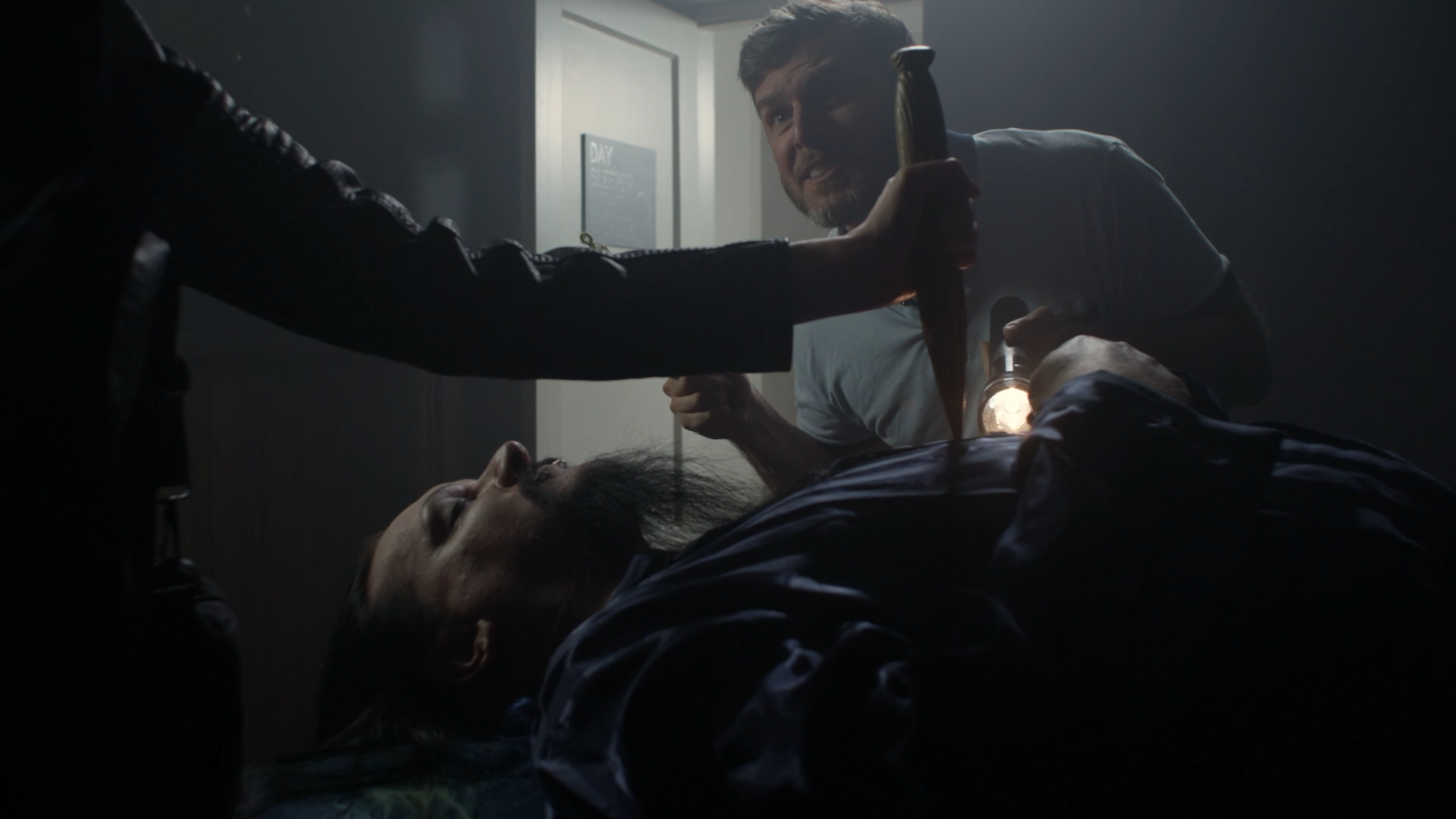 A man with long dark hair and a beard lying on a bed, while another man with short dark hair and a beard is holding a hammer and a flashlight in a dimly lit room.