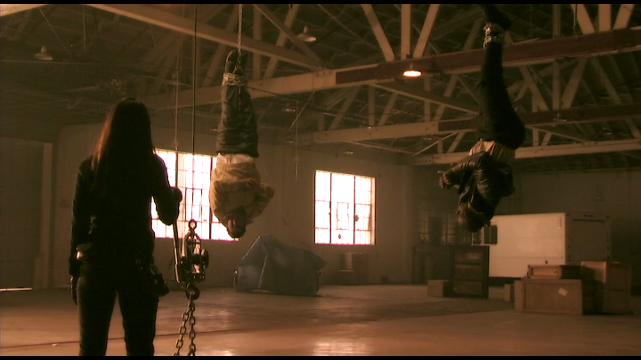 Three people in an industrial warehouse, with two hanging upside down and one standing, during a scene from a movie or photoshoot.
