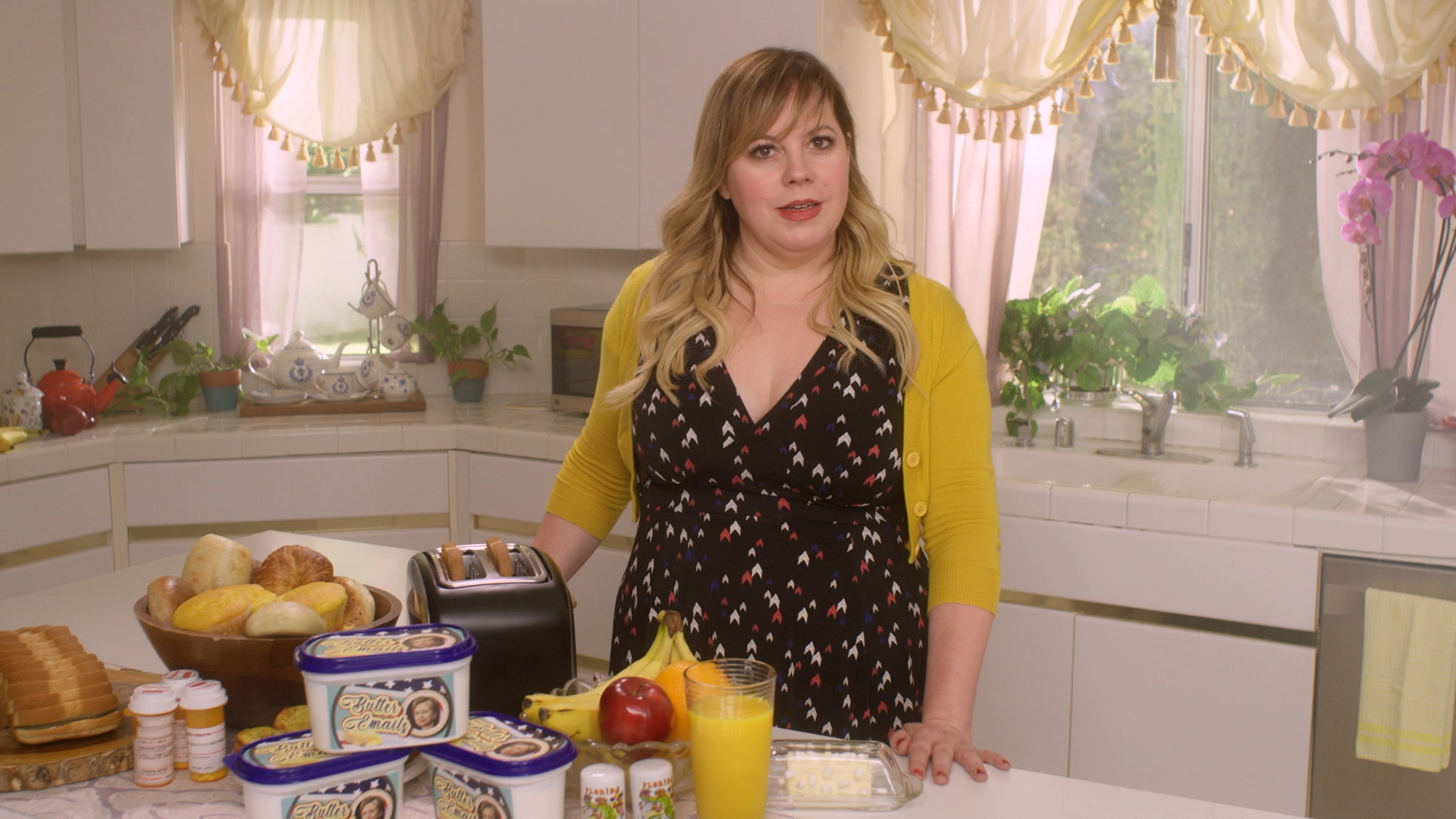 A woman in a black dress with small multicolored patterns and a yellow cardigan standing in a kitchen with breakfast foods on the counter. The kitchen has white cabinets, a window with pink curtains, and pink orchids.