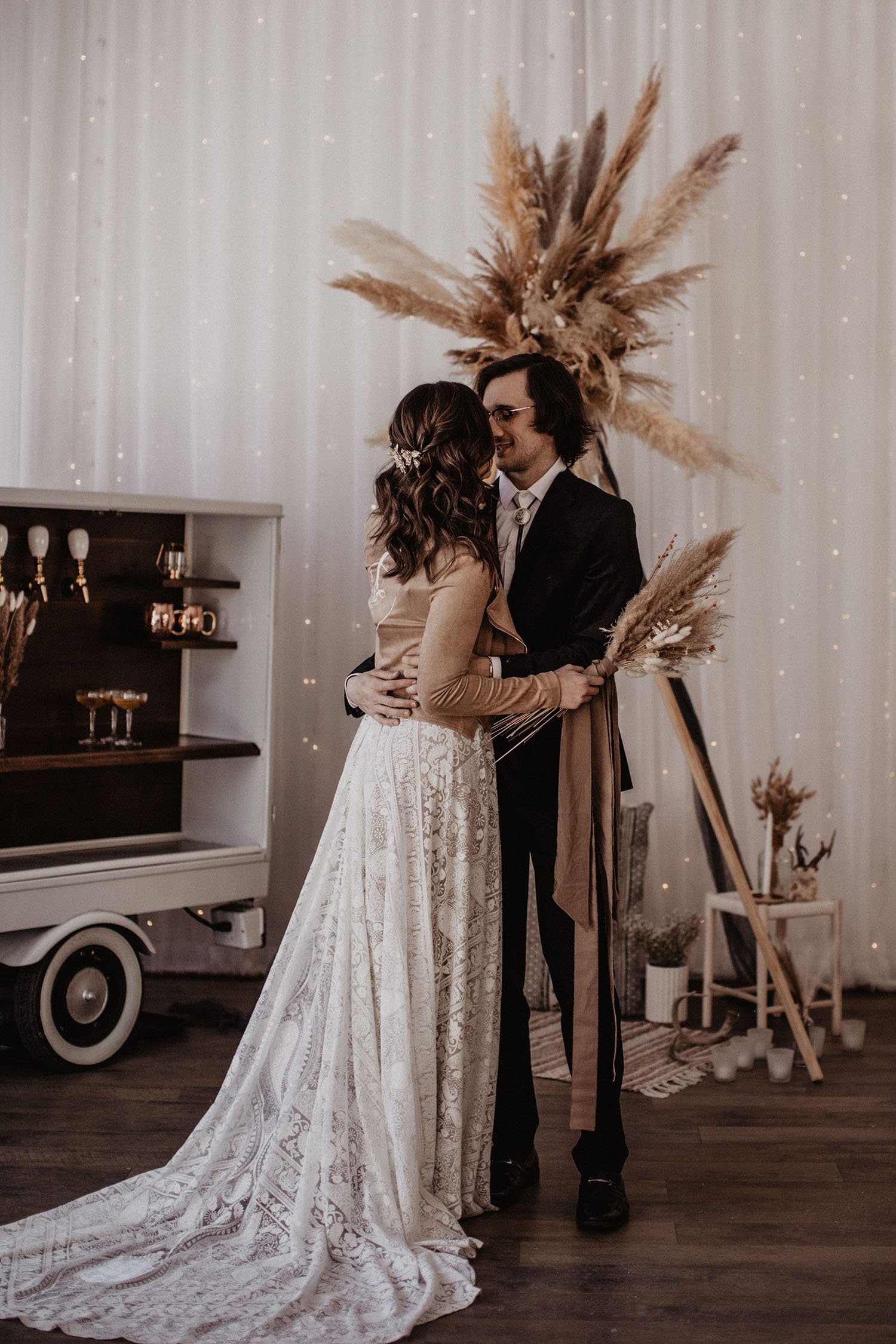  A bride wearing the Rue de Seine kyara wedding dress with a brown leather jacket holding a bridal bouquet of dried florals. She is standing facing her groom with their arms around each others waist. They are standing in front of a boho wearing arbor