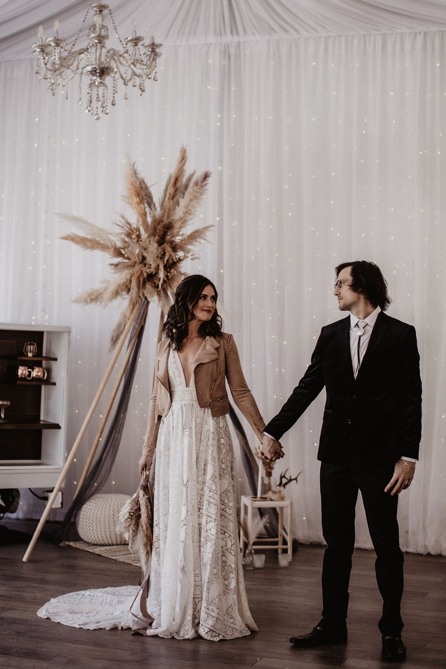  A bride wearing Rue de Seine Kyara wedding dress, which is a deep-v banded waist bold lace dress with a dramatic train. She is also wearing a brown leather jacket and holding a bouquet of pampas grass in one hand and the grooms hand in the other. Th