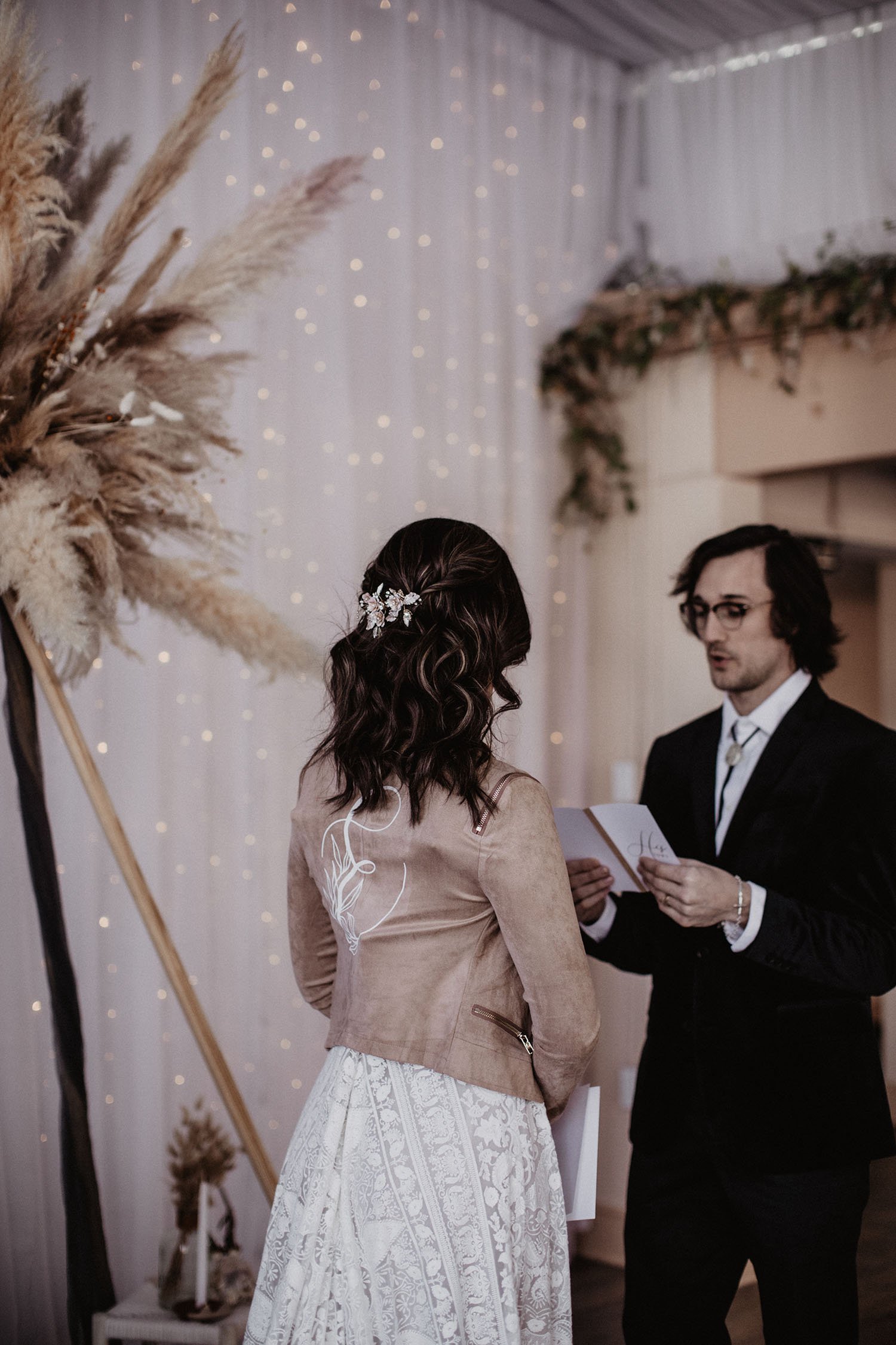  A bride wearing Rue de Seine Kyara wedding dress and a brown suede jacket with a monogram ‘E’ and florals painted on the back. Her hair is half up in soft curls with a hair comb of gold and ivory. She is standing with her back to the camera facing t