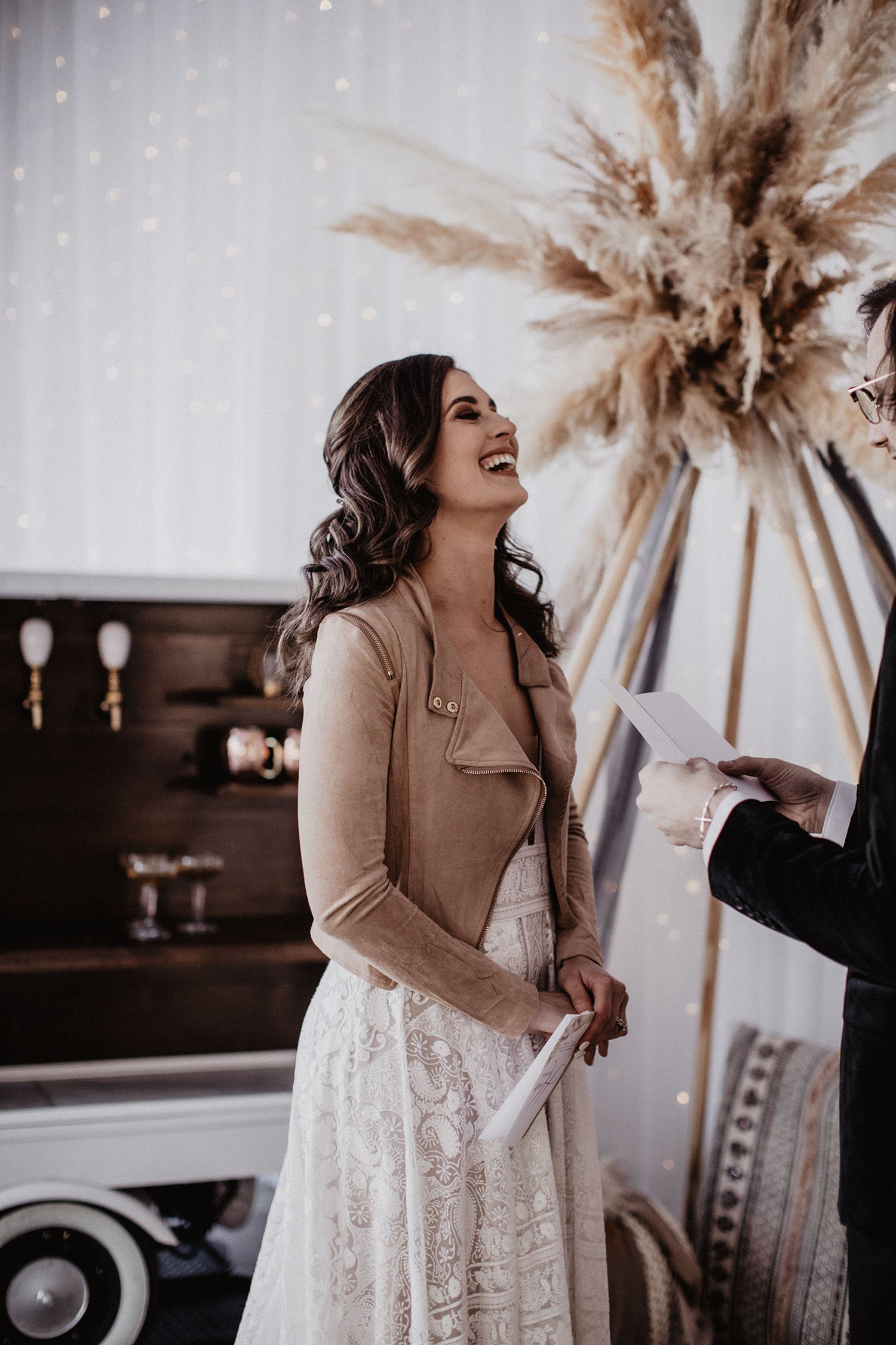 A bride wearing Rue de Seine Kyara, which is a bold lace wedding dress with a thick banded waist and deep-v neckline. She is wearing a brown suede leather jacket and her hair is tilted back laughing. Her hair is dark brown and styled half up in soft