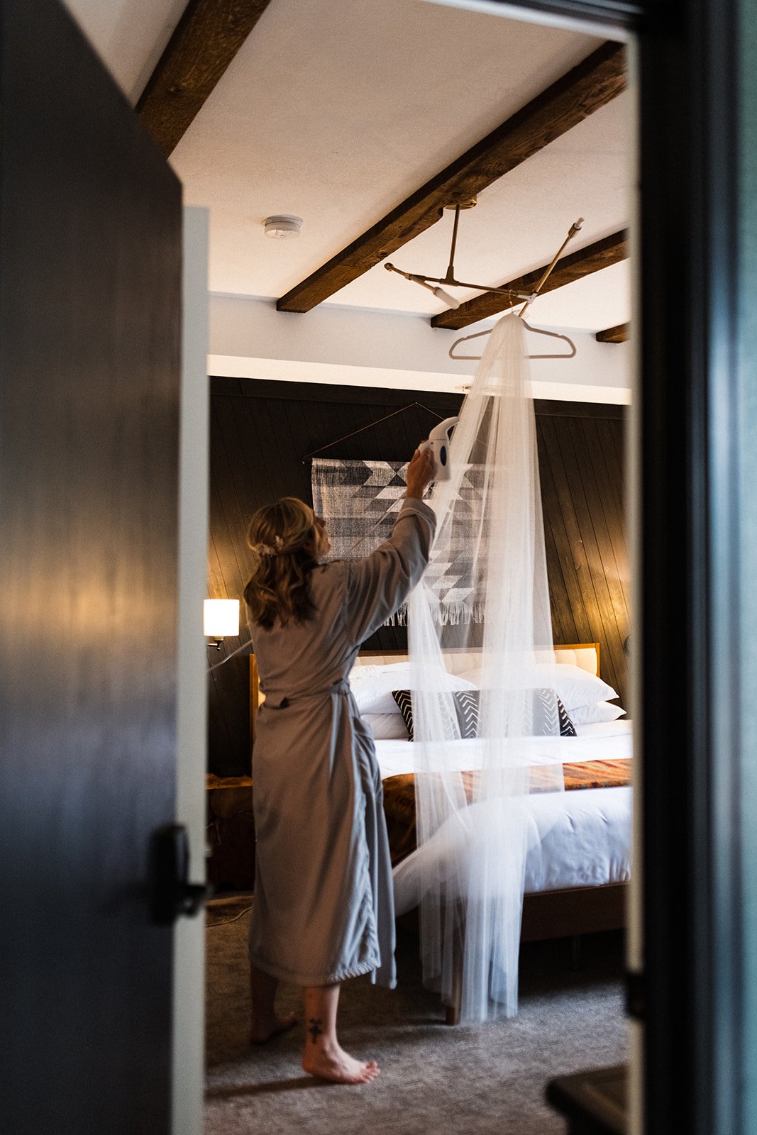  Photograph of a bride getting ready for her big day while wearing a plush grey robe and steaming her Sara Gabriel veil in the modern bedroom of a mountain cabin. 