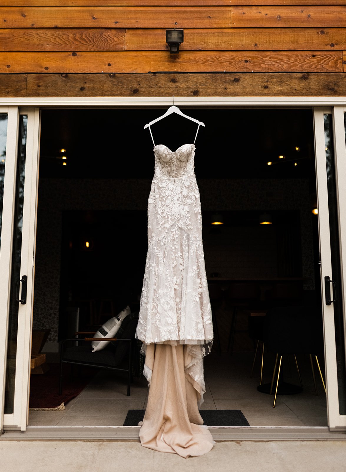  Photograph of the Made With Love Penny wedding dress hanging on the trim of large double doors leading out onto a balcony. It’s a strapless fitted wedding dress with 3D lace appliqués and a nude mesh lining with a long train. 