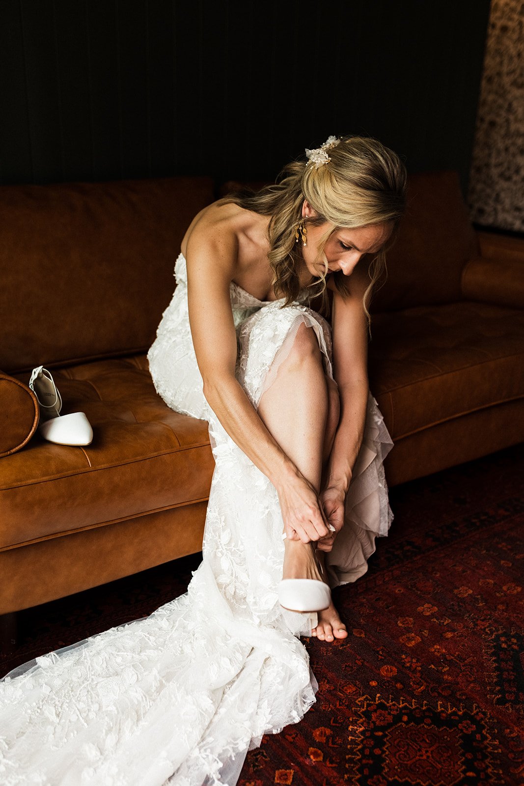  Photograph of a bride wearing the Made With Love Penny wedding dress sitting on a leather tufted sofa. She is bending over to clasp the ankle strap on her white d'orsay heels. Her hair is in soft curls with two tendrils falling around her face. 