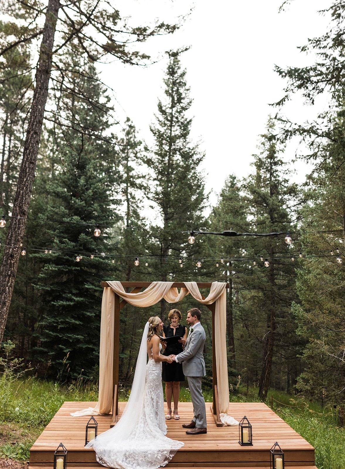  Photograph of a bride and groom standing at an alter in the woods as they exchanging vows for their mountain elopement. The bride is wearing Made With Love Penny wedding dress and a Sara Gabriel tulle veil. They are holding hands as their officiant 