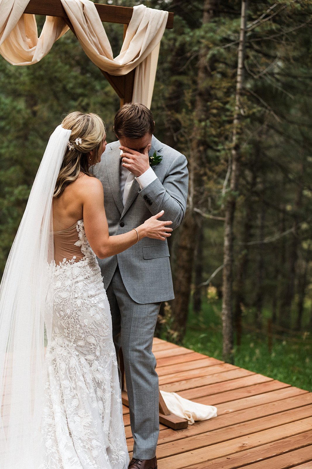  Photograph of a bride wearing Made With Love penny wedding dress, Sara Gabriel veil, and floral hair pieces. She is reaching out to the groom who is wiping tears from his eyes as they exchange vows for their intimate mountain elopement in the woods.