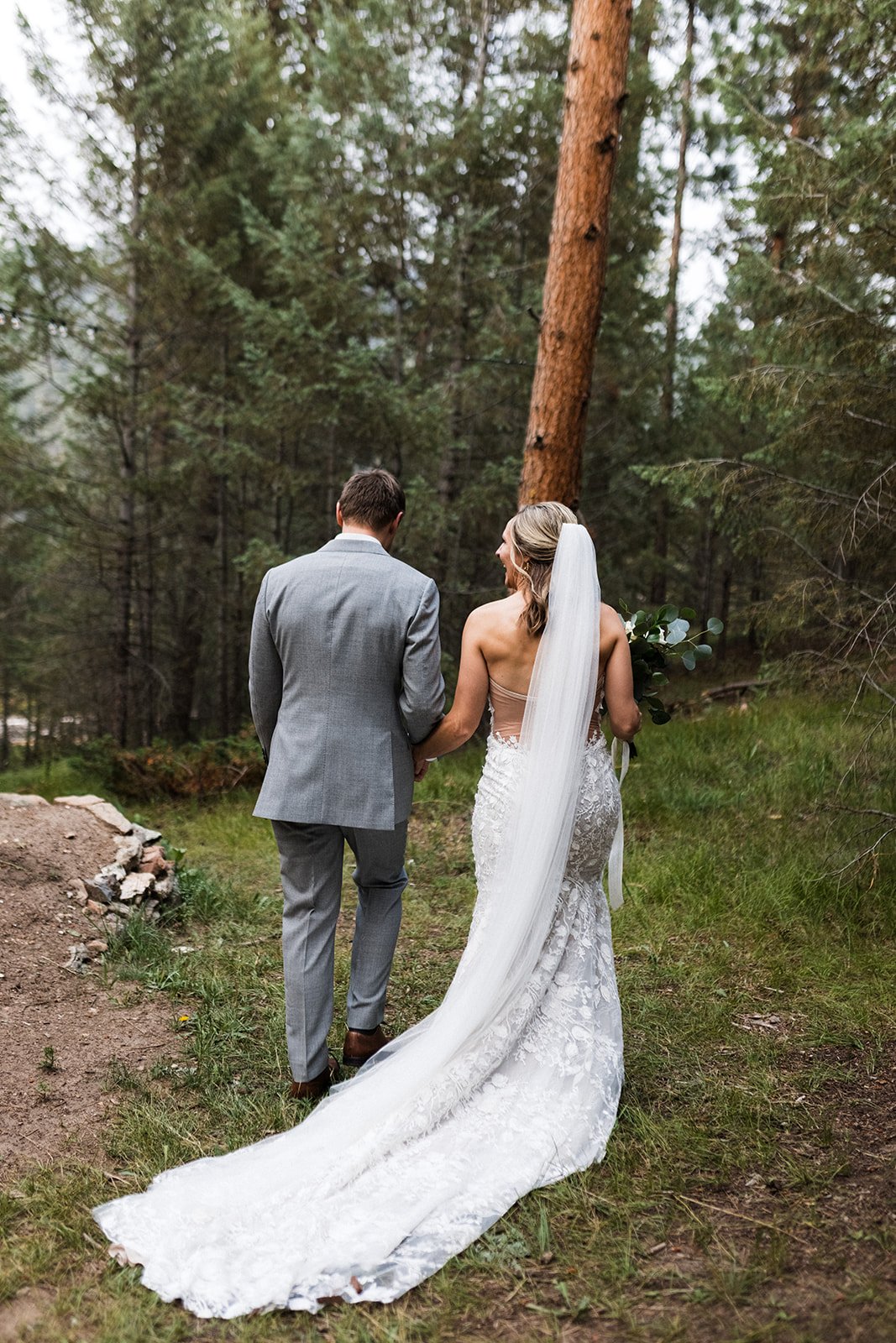  Photograph of a bride and groom walking away after exchanging vows for their intimate woodland elopement in the mountains. The bride is wearing the Made With Love Penny dress, which has a long lace train that is trailing her bind her along with her 