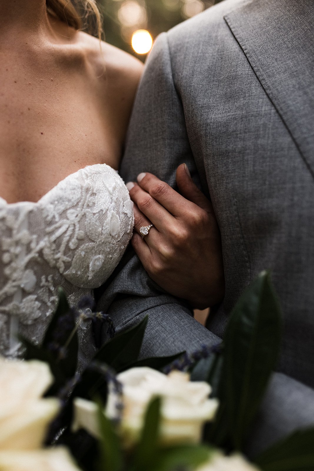  An up close photograph of a bride wearing Made With Love Penny wedding dress, which has a strapless bustier bodice and 3D lace appliqués over a nude lining. She is wearing an oval cute diamond engagement ring with her hand looped through the grooms 