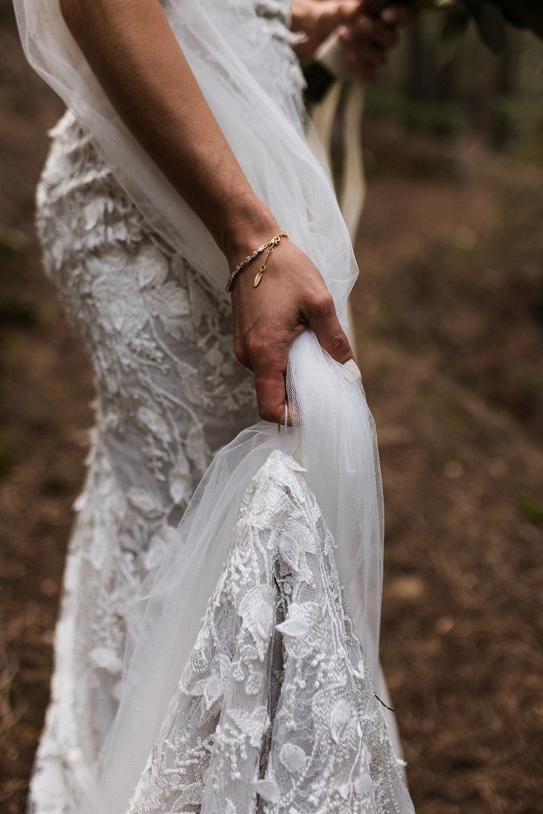  A photograph of the waist down of a bride wearing Made With Love Penny wedding dress and a Sara Gabriel veil. She has the trail of her wedding dress and her veil clasped in her left hand. Her nails are painted a soft pink and she is wearing a gold a