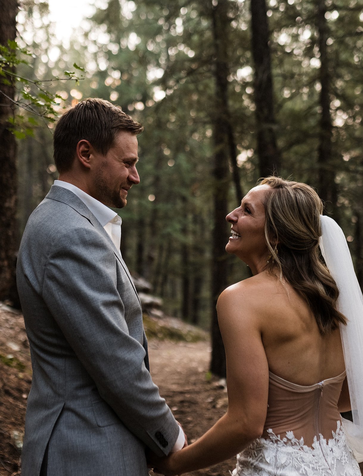  Photograph of a bride and groom holding hands facing each other and laughing in the woods after their mountain elopement in the forest. The bride is wearing Made With Love Penny, which is a nude mesh low back and 3D lace appliqués. Her hair is half 