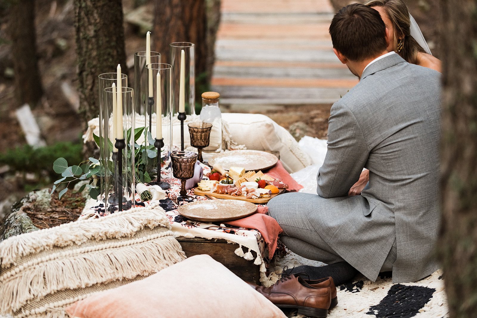  Photograph of a bride and groom having a boho woodland picnic after their mountain elopement in the forest. They are seating on Moroccan poufs on the ground at a low dining table covered in tasseled linens, black taper candles, and purple vintage go