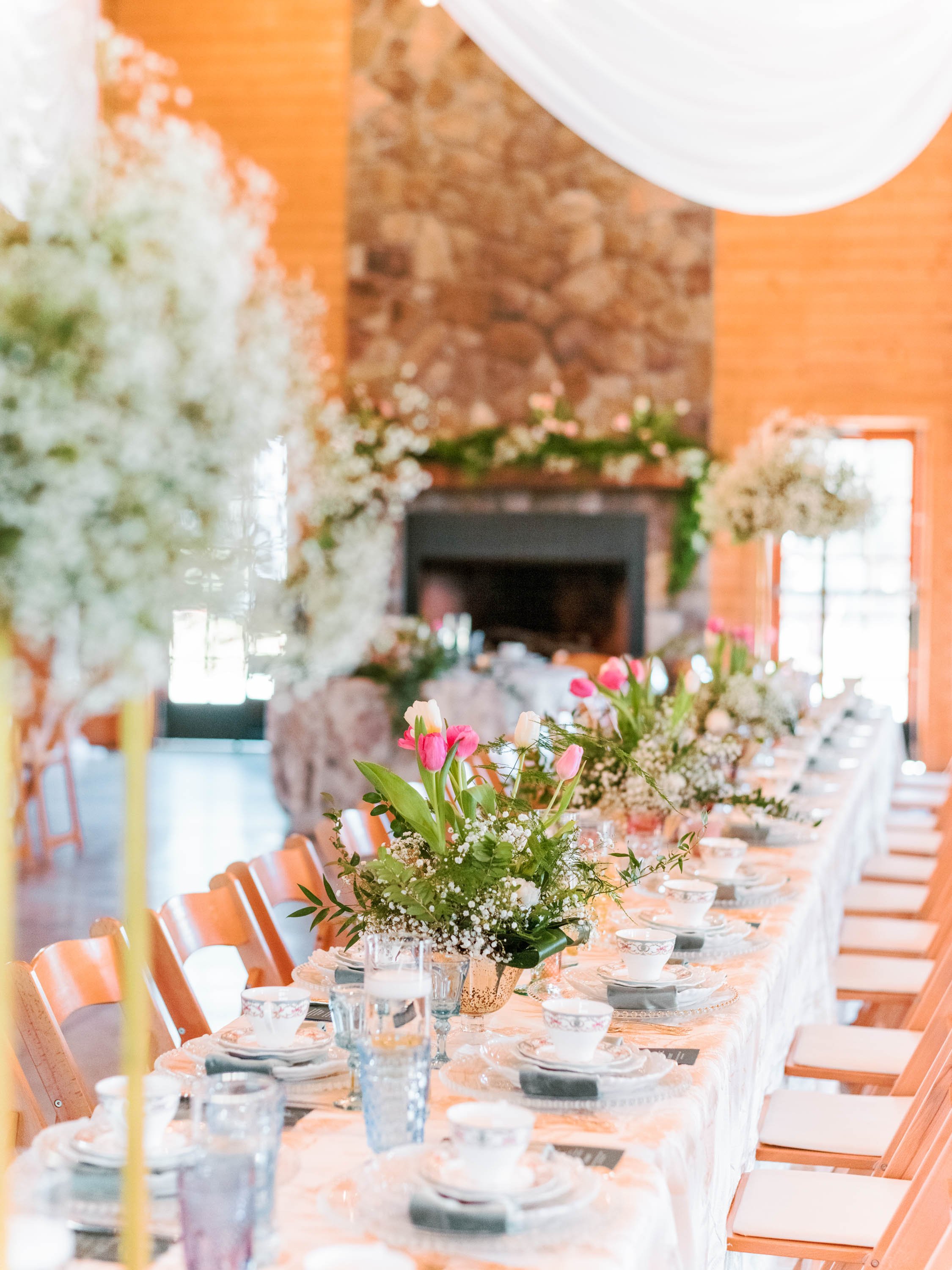  A vibrant spring tablescape at Oakland Farm wedding venue near Raleigh, North Carolina. The table is covered in white linens, vintage place settings with teacups, and is lined with center pieces of pink tulips, ferns, and babies breath. There is a s