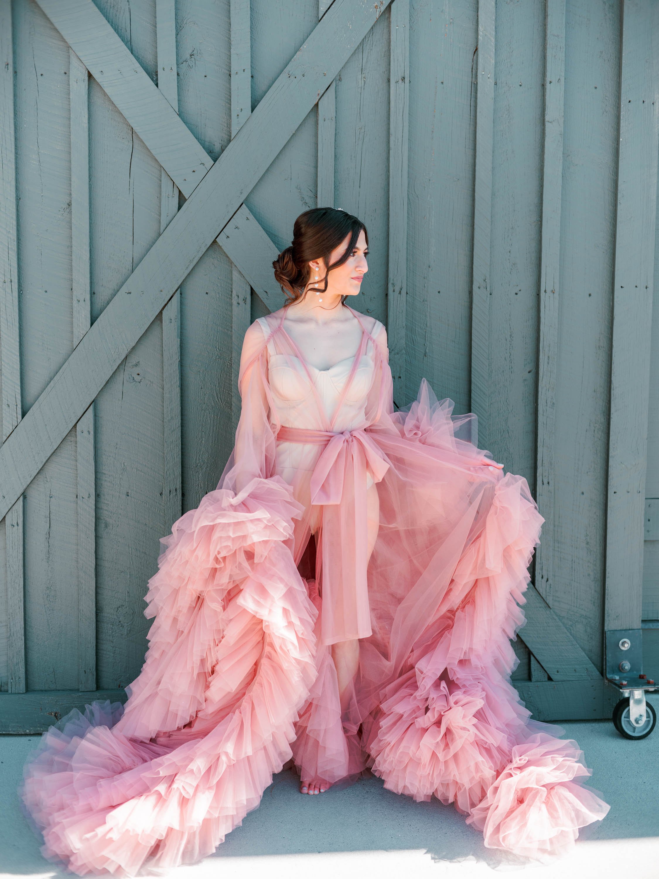  A bride standing in front of a blue barn door wearing a sheer pink tulle bridal robe with full cascading ruffles. Her hair is dark brown and styled back in a low chignon with tendrils curled around her face, and tiered pearl drop earrings as she loo