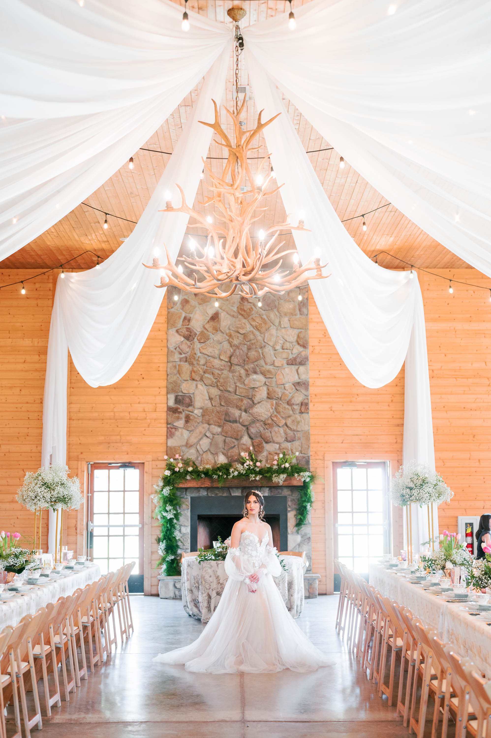  A bride wearing Chic Nostalgia Ayla standing in front of a fireplace in the reception hall at Oakland Farms near Raleigh, North Carolina. The bridal gown has a strapless sweetheart bodice adorned with floral lace appliqués and a full tulle skirt wit