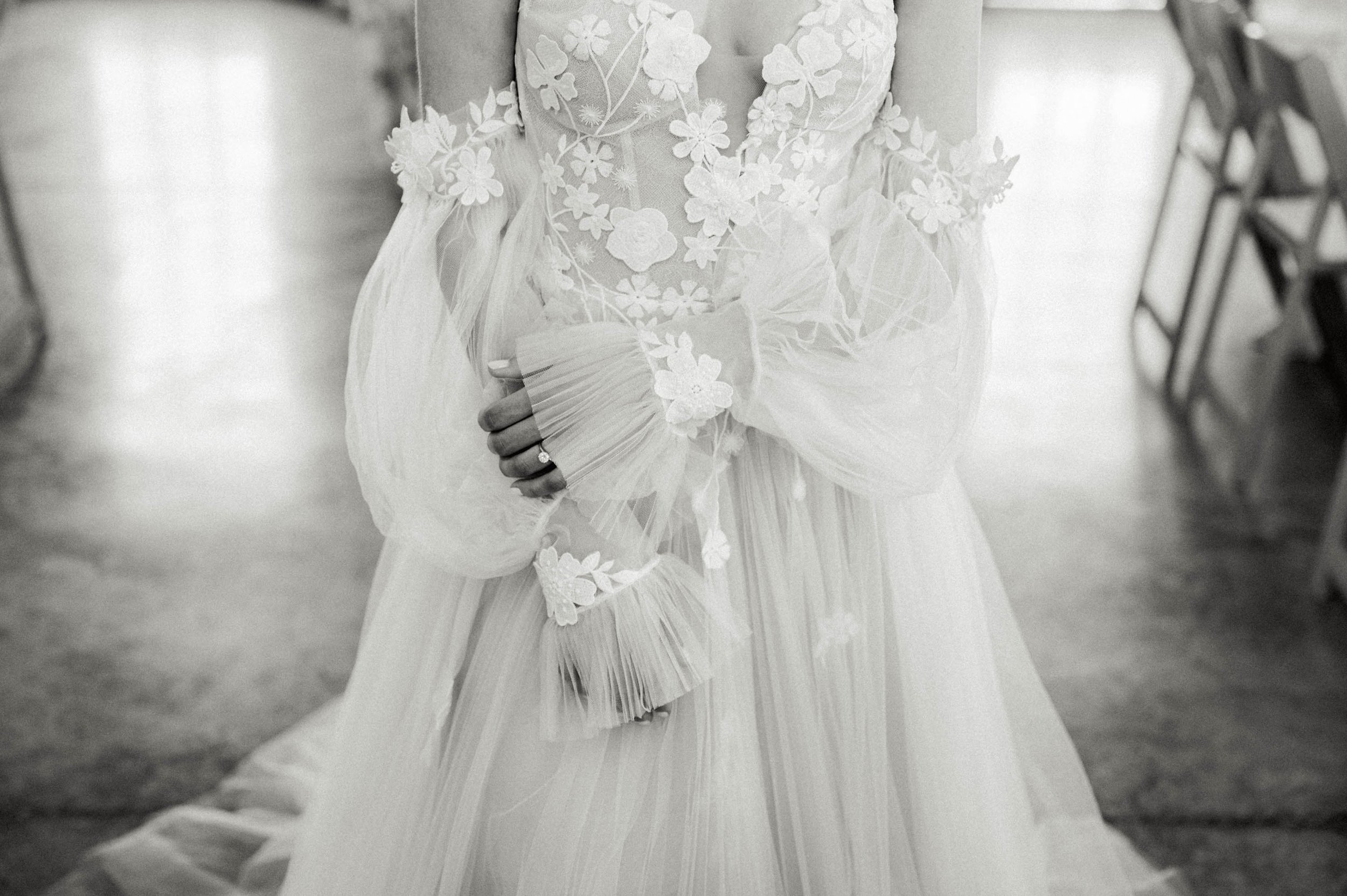  A black and white photograph of a bride in Chic Nostalgia Ayla wedding dress, which is a bustier bodice with a sheer deep v covered in large floral 3D lace. The wedding dress has a full tulle skirt and an off the shoulder puff tulle sleeves with ruf