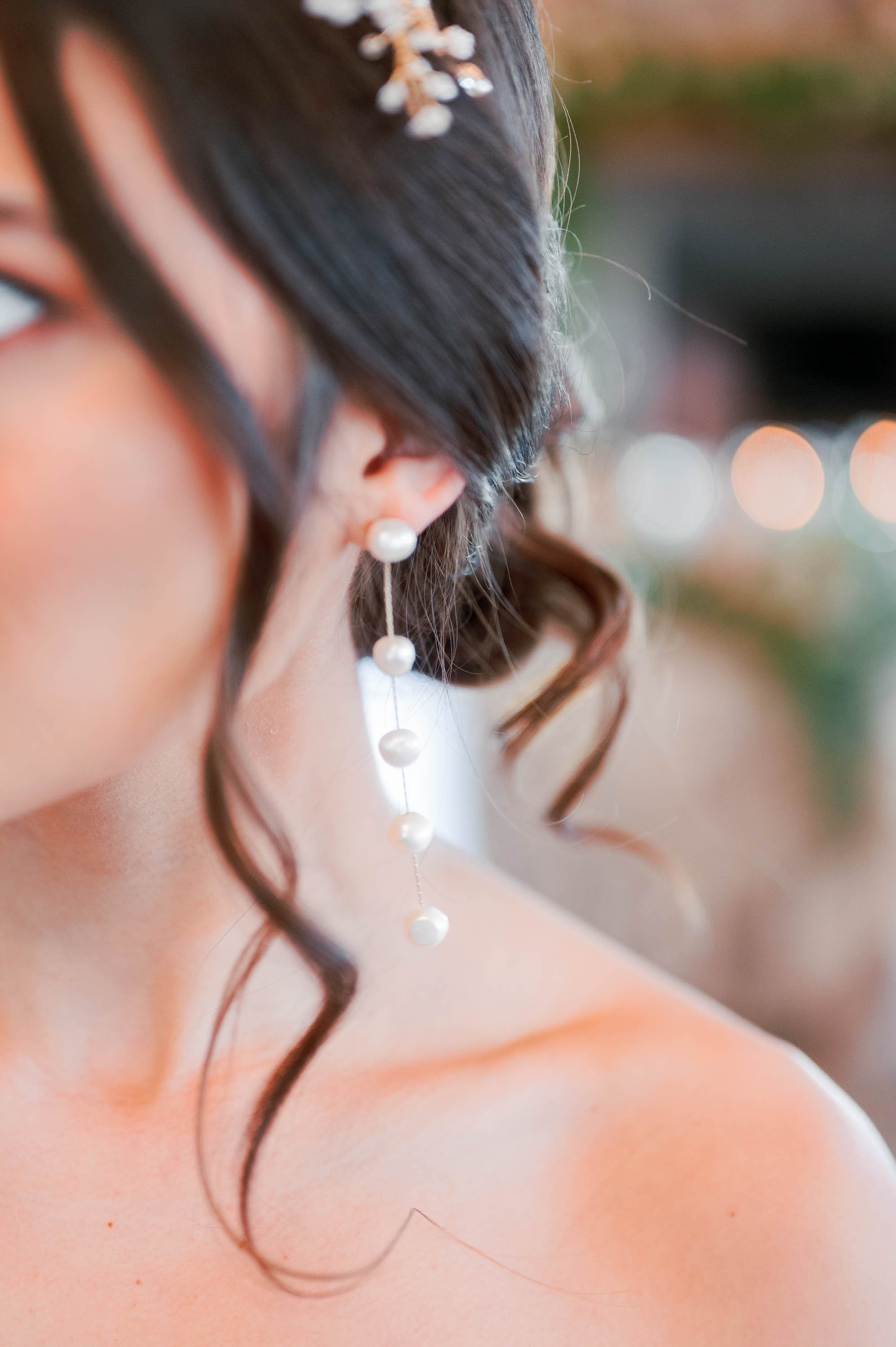  A close up of a bride in a strapless wedding dress with her dark brown hair in a low chignon and soft tendrils curled around her face. She is wearing tiered pearl drop earrings and there is a soft bokeh in the background.  