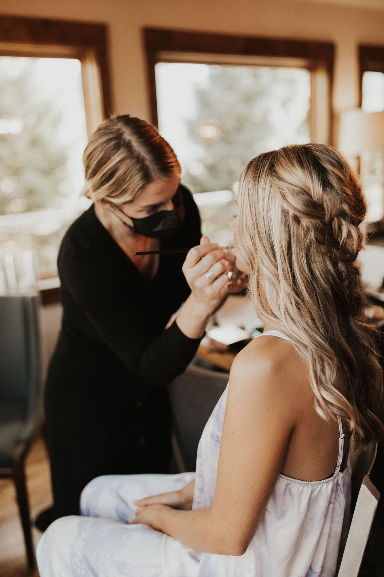  photograph of a makeup artist getting a bride ready for her big day. The bride has her long blonde hair done half up with loose braids and soft curls as the HAMU paints her lips. 