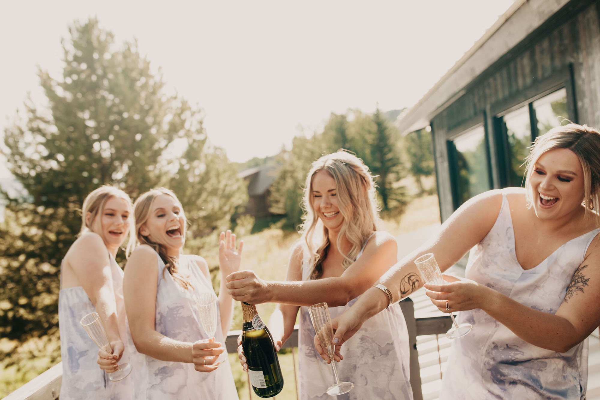  Photograph of a bridal party popping a bottle of champagne on a deck in the mountains of Crested Butte. They are all laughing and wearing matching silk tie dye pajamas. 