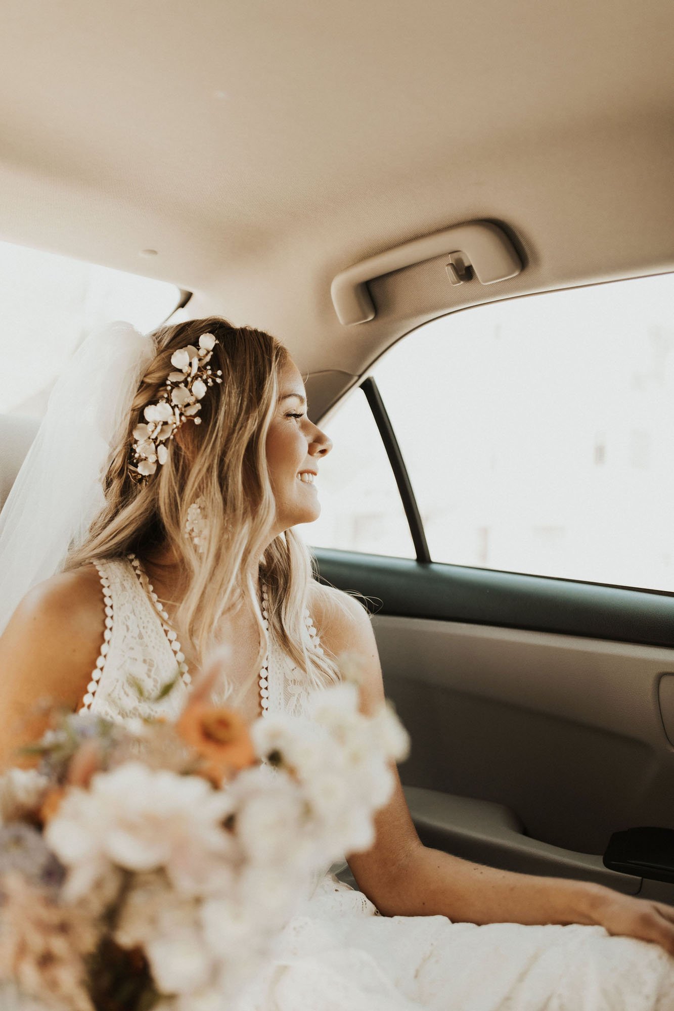  Photograph of a bride wearing Rish Rio wedding dress in the back seat of a car on the way to her ceremony location in Crested Butte. She is smiling looking out the window with a floral comb in her hair and a tulle veil on top of her head. You can se