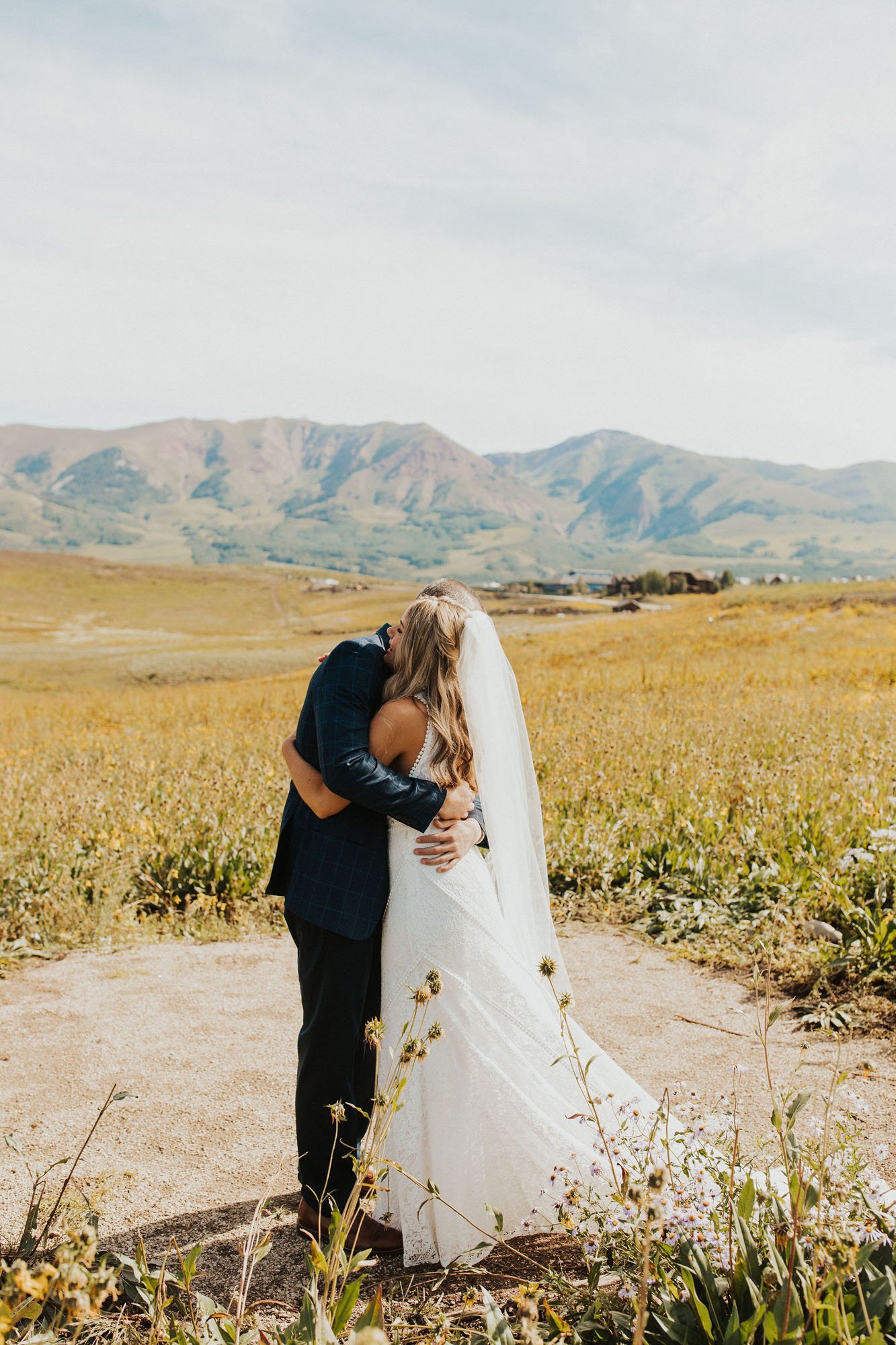  Photograph of a bride wearing Rish Rio Wedding dress and hugging the groom as they have their first look in a field of wildflowers on a sunny day in Crested Butte, Colorado. They are embracing with their hands around each other’s waist, with the bri