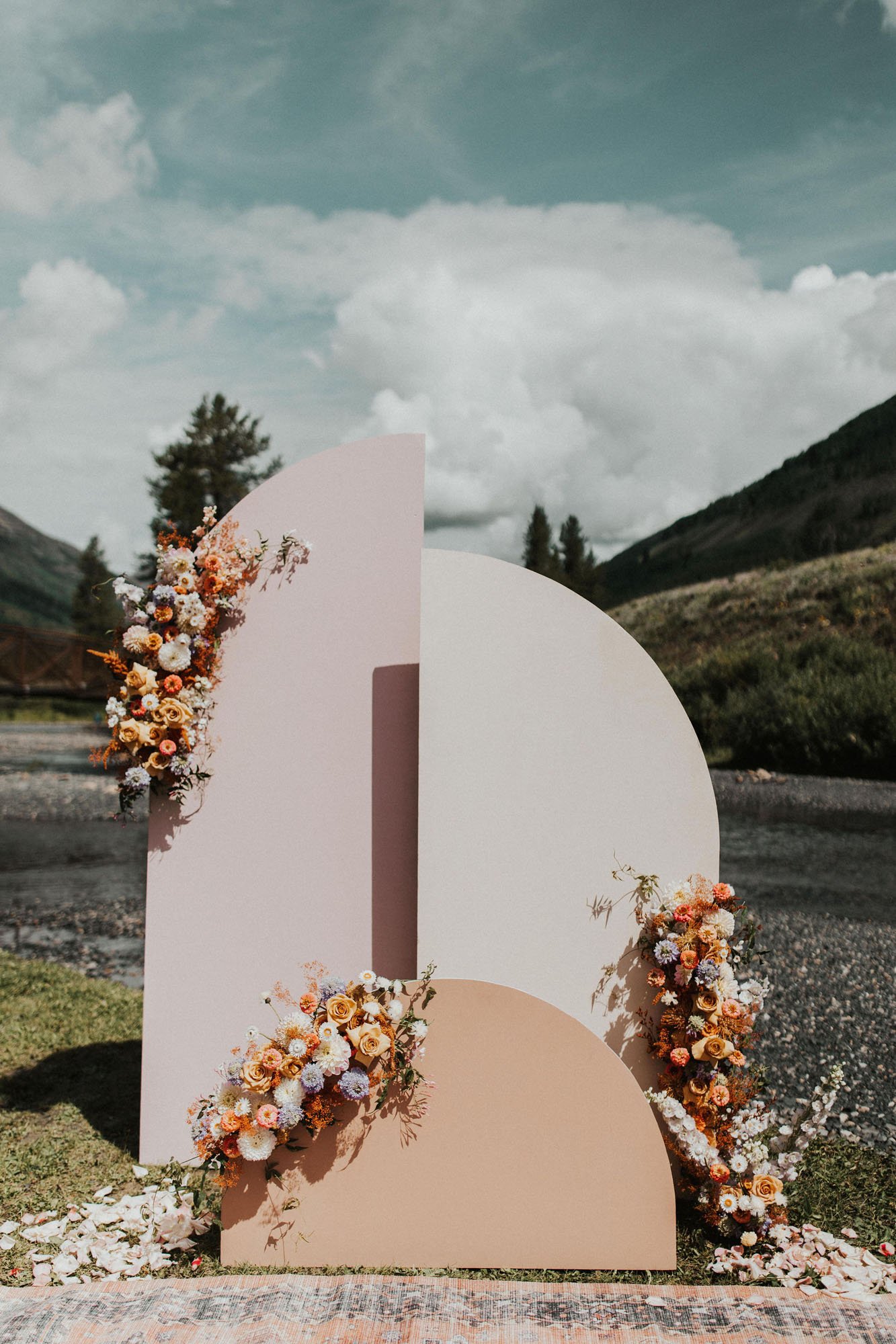  Photograph of a modern geometric ceremony backdrop in front of a creek in Crested Butte, Colorado. The backdrop is made of semi-arches of varying heights and shades of beige and pink and is decorated with florals in white, orange, beige, purple, and