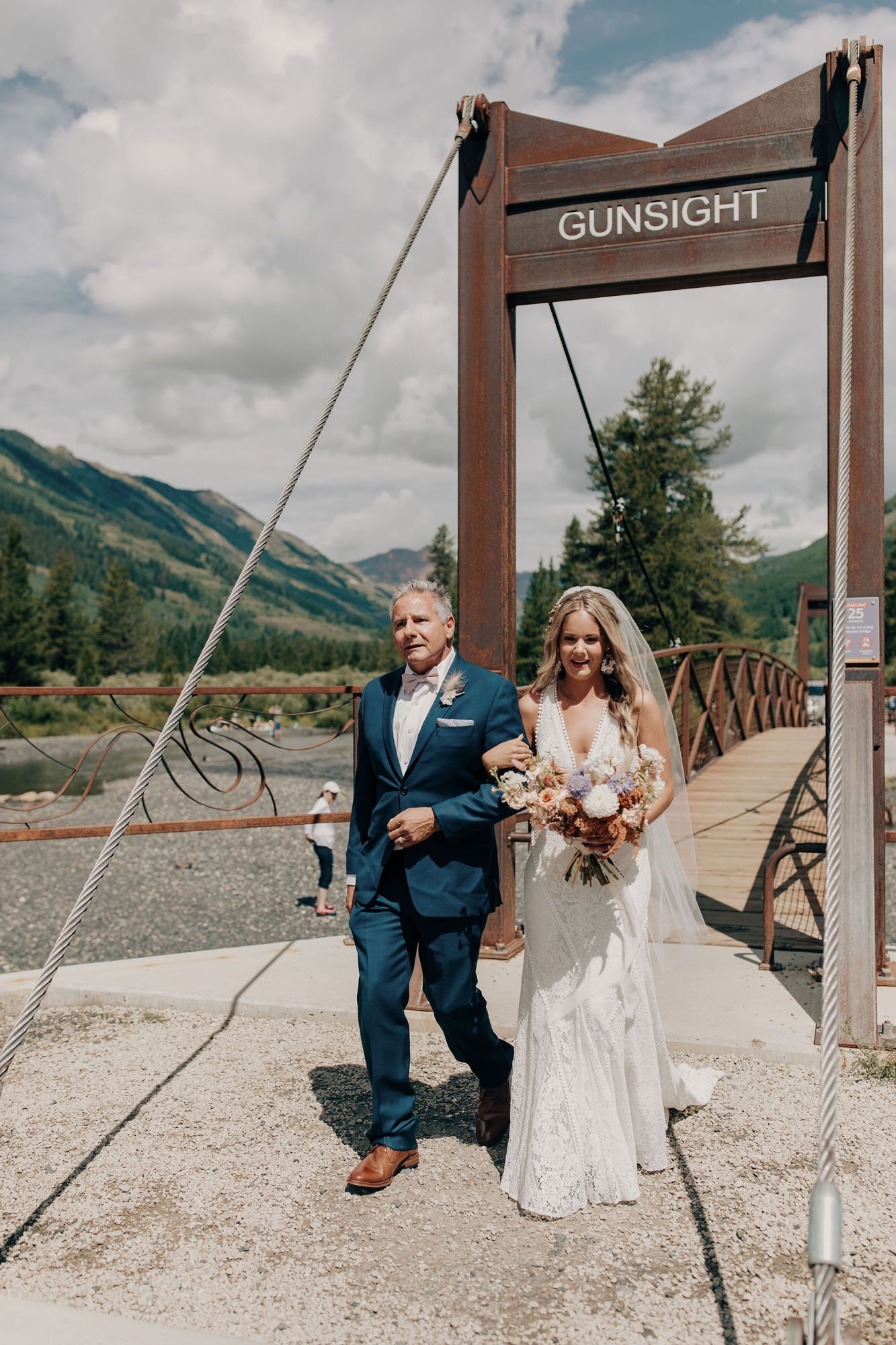  Photograph of a bride wearing the Rish Rio wedding dress walking across slingshot bridge in crested butte escorted by her father. She is wearing a Sara Gabriel wedding veil and carrying a bridal bouquet of white, blush, beige, and purple flowers.  