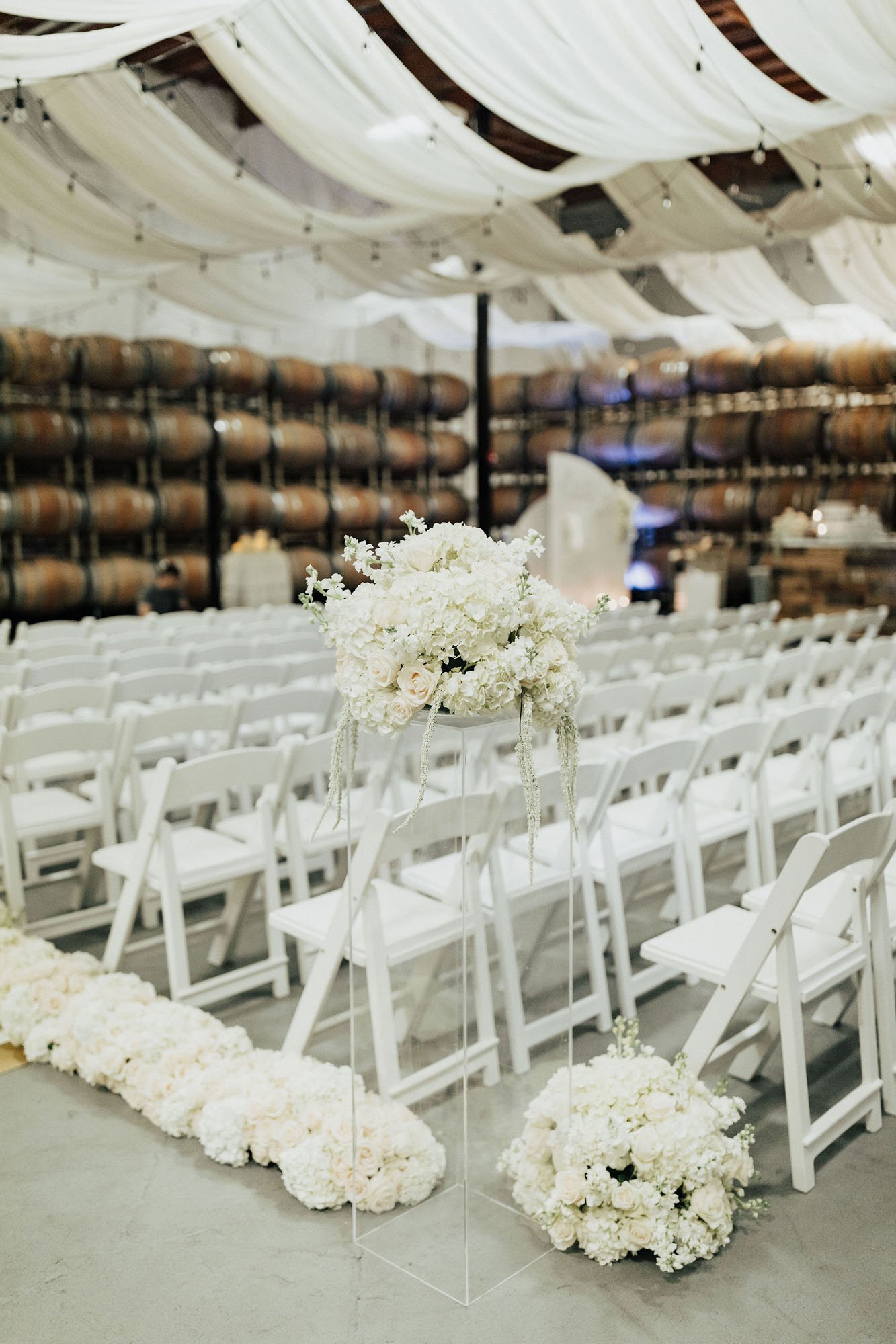  Photograph of a ceremony setup in a PNW vineyardr in Woodinville, WA. The walls are lined with oak wine barrels, the ceilings are draped with white silk, and there are decorative white wood folding chairs and white rose and hydrangea floral arrangem