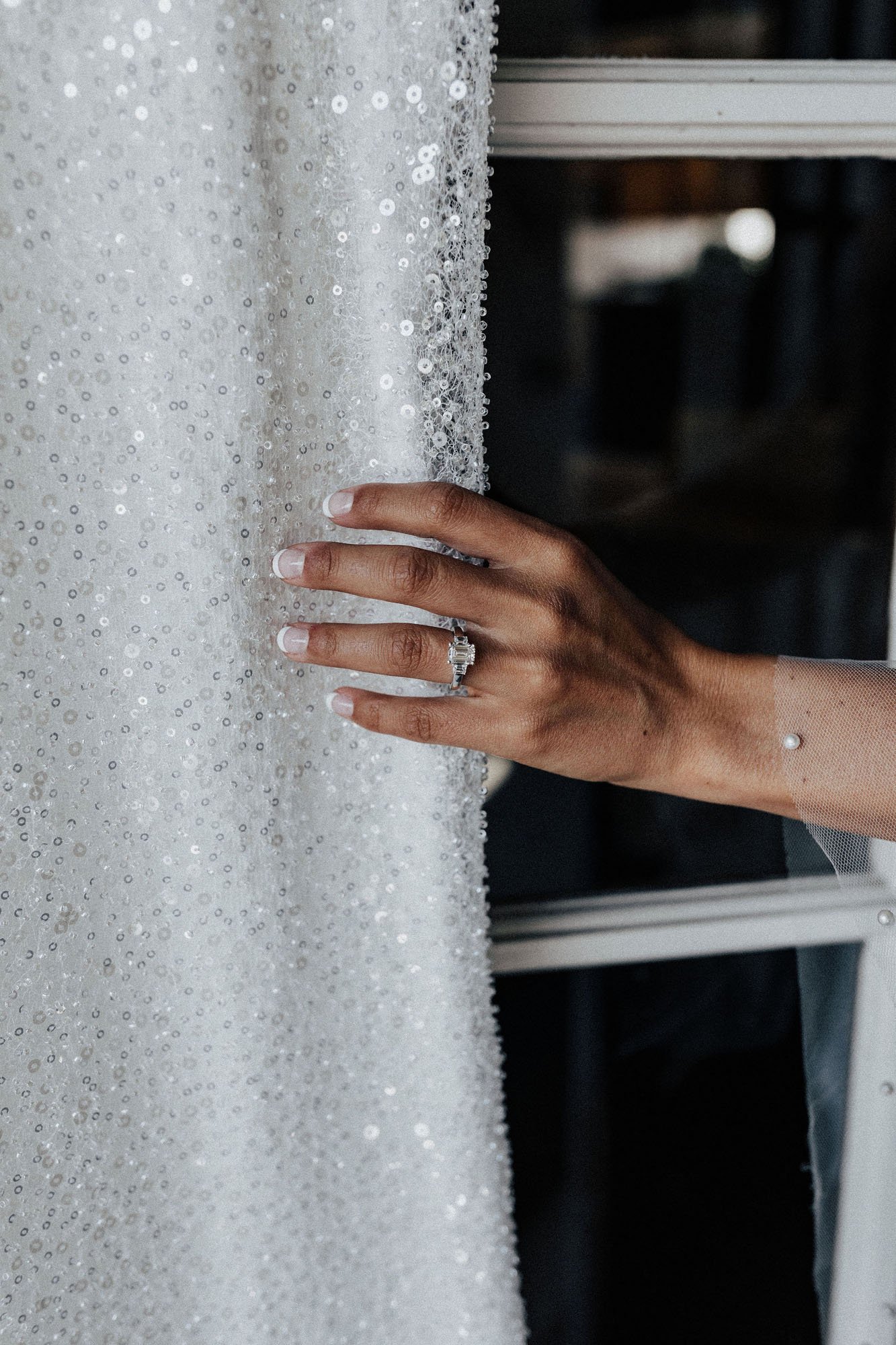  up close photograph of a bride’s hand holding the skirt of the Made With Love ‘Lola’ beaded wedding dress. Her nails have a french Manicure and she is wearing an emerald cut engagement ring and a sheer tulle robe with pearls. 