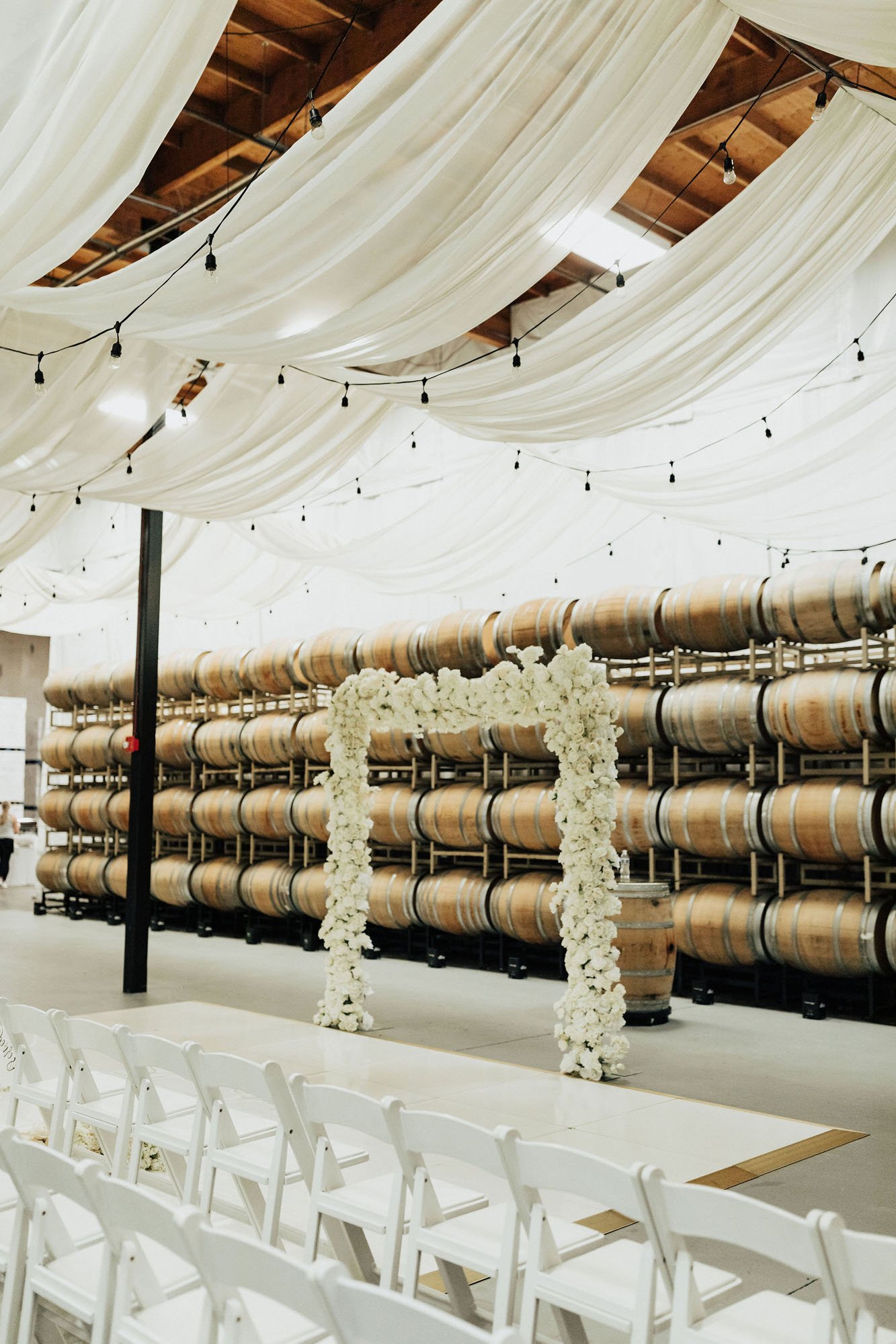  Photograph of a modern ceremony arbor in a vineyard in Washington. There is a large white floral arbor covered in white roses and hydrangeas in front of a wall of oak barrels for storing wine. The ceilings are draped with white silk and bistro light