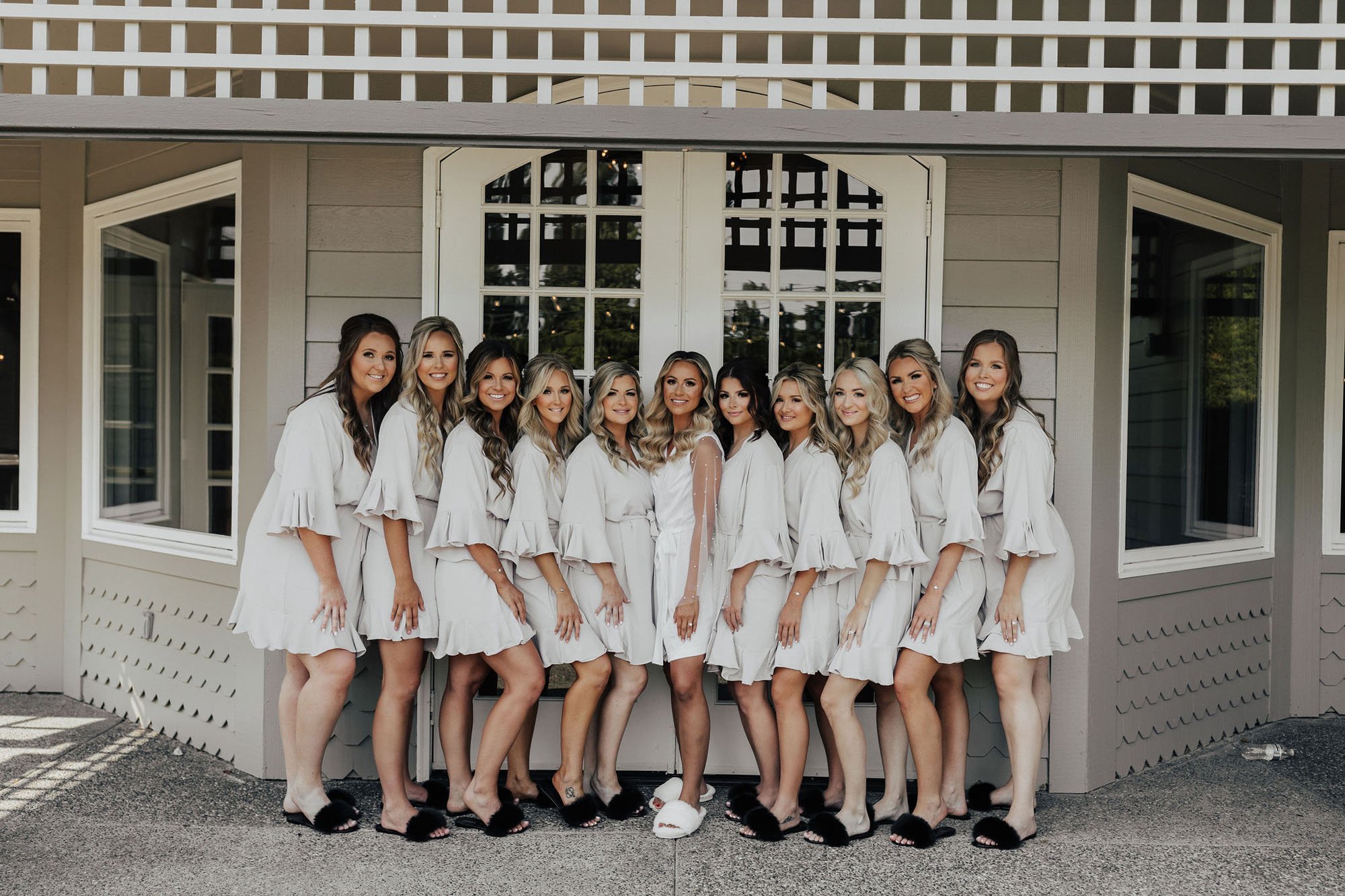  A photograph of a bridal party with ten bridesmaids. The bride is in the center of the group and they are all standing under an awning in front of large glass double doors. They are wearing short ruffle robes and fuzzy slippers as they get ready for