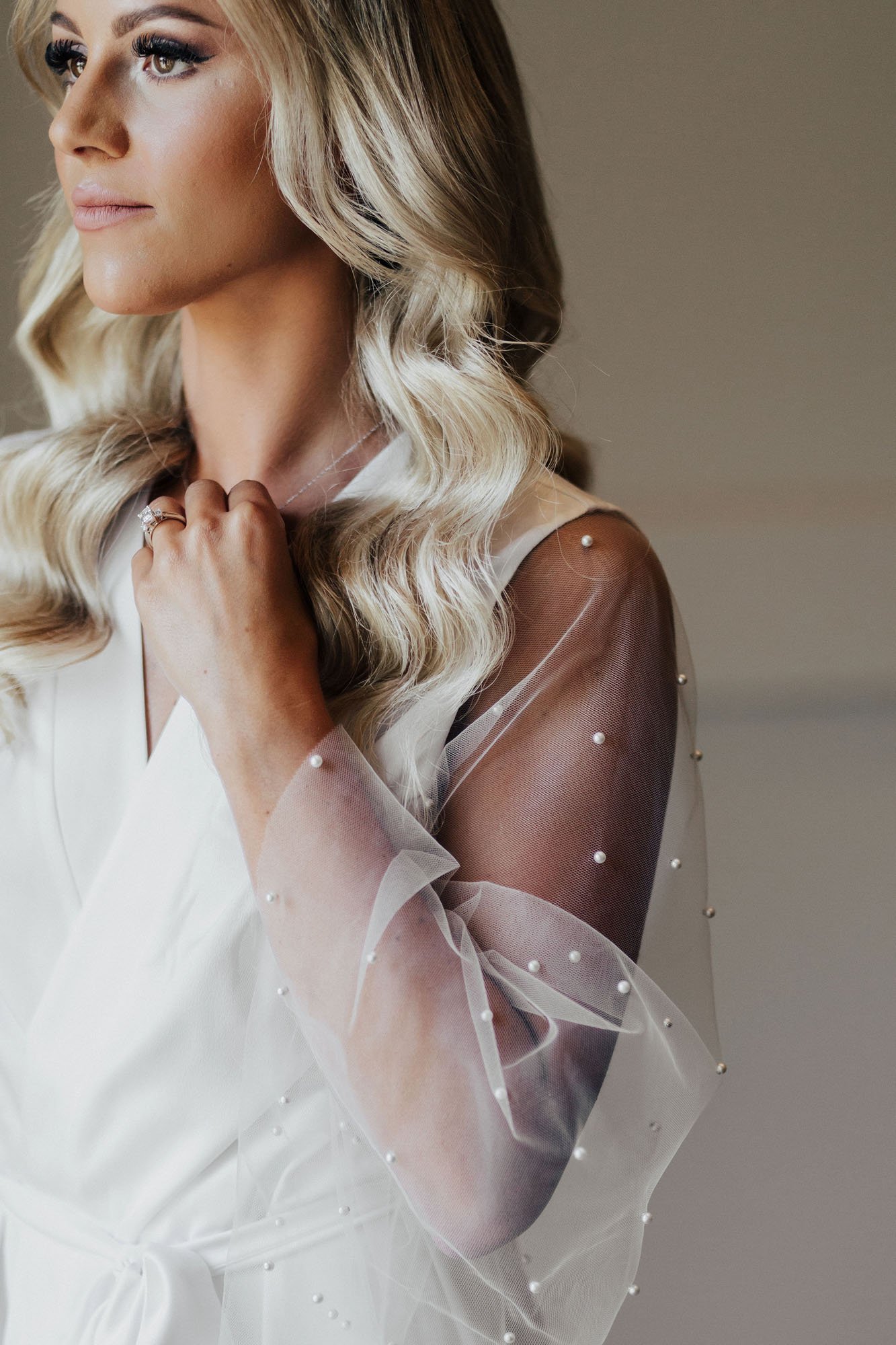  Photograph of a bride with long blonde hair in soft curls. She is wearing a white robe with sheer tulle sleeves flocked in pearls, and is looking off in the distance. She has her left hand help up to her neck holding her necklace, and is wearing an 