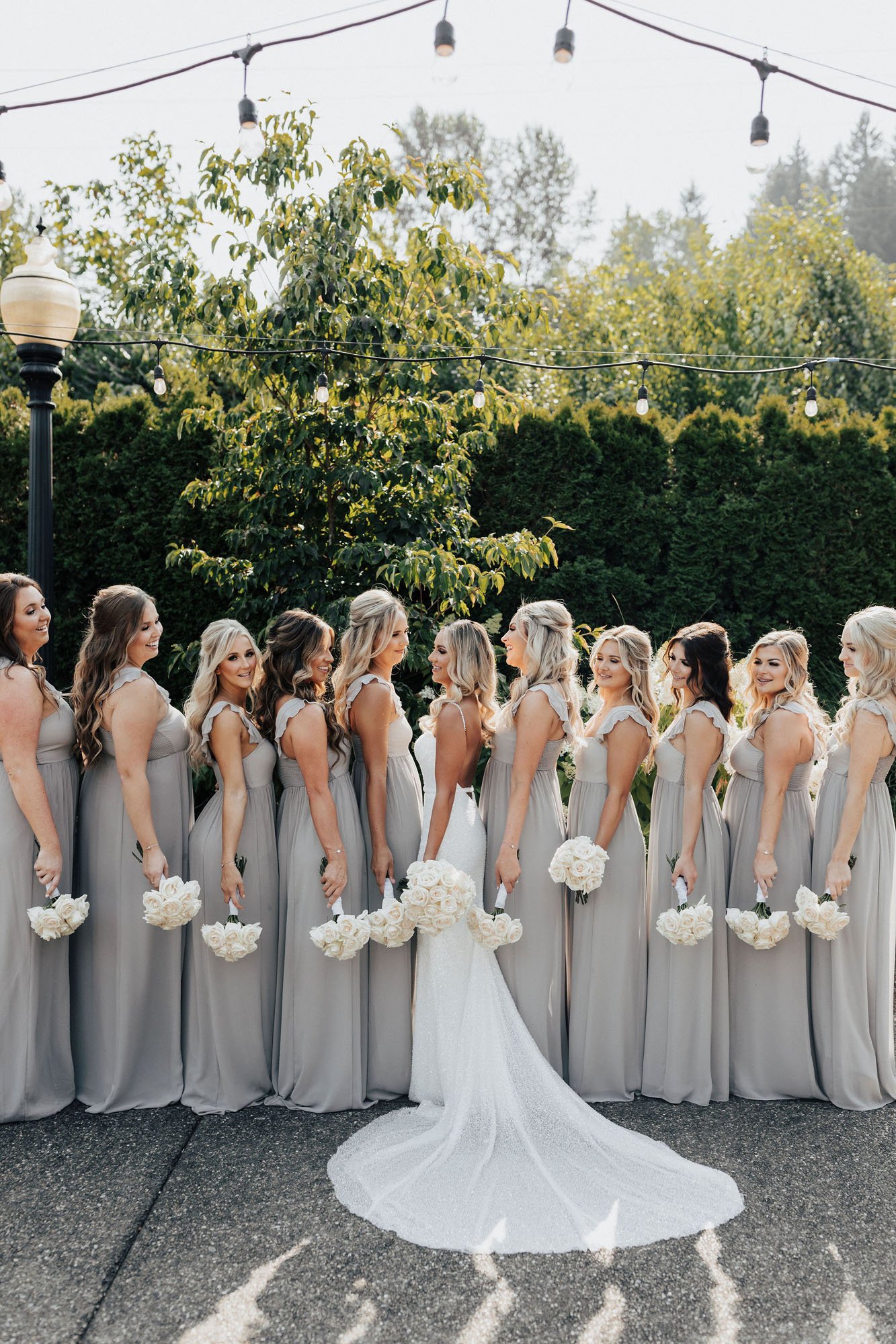  Photograph of a bridal party with ten bridesmaids. The bride is standing in the center of the group wearing the Made With Love Lola wedding dress, which has a low back and is covered in sequins. The bridesmaids are wearing grey chiffon floor length 