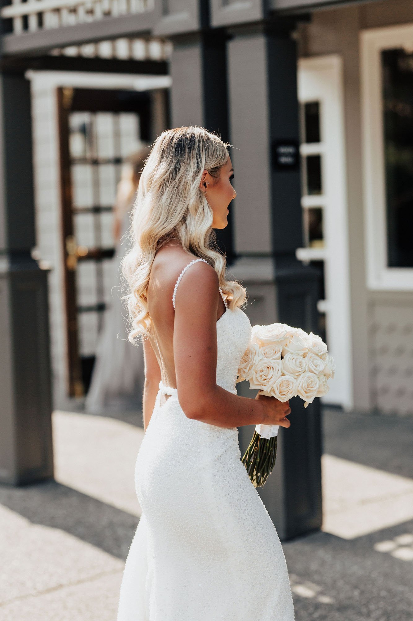  Photograph of the back of a bride wearing Made With Love Lola wedding dress and walking while holding a bouquet of white roses. She has long soft curly blonde hair and a tan. 