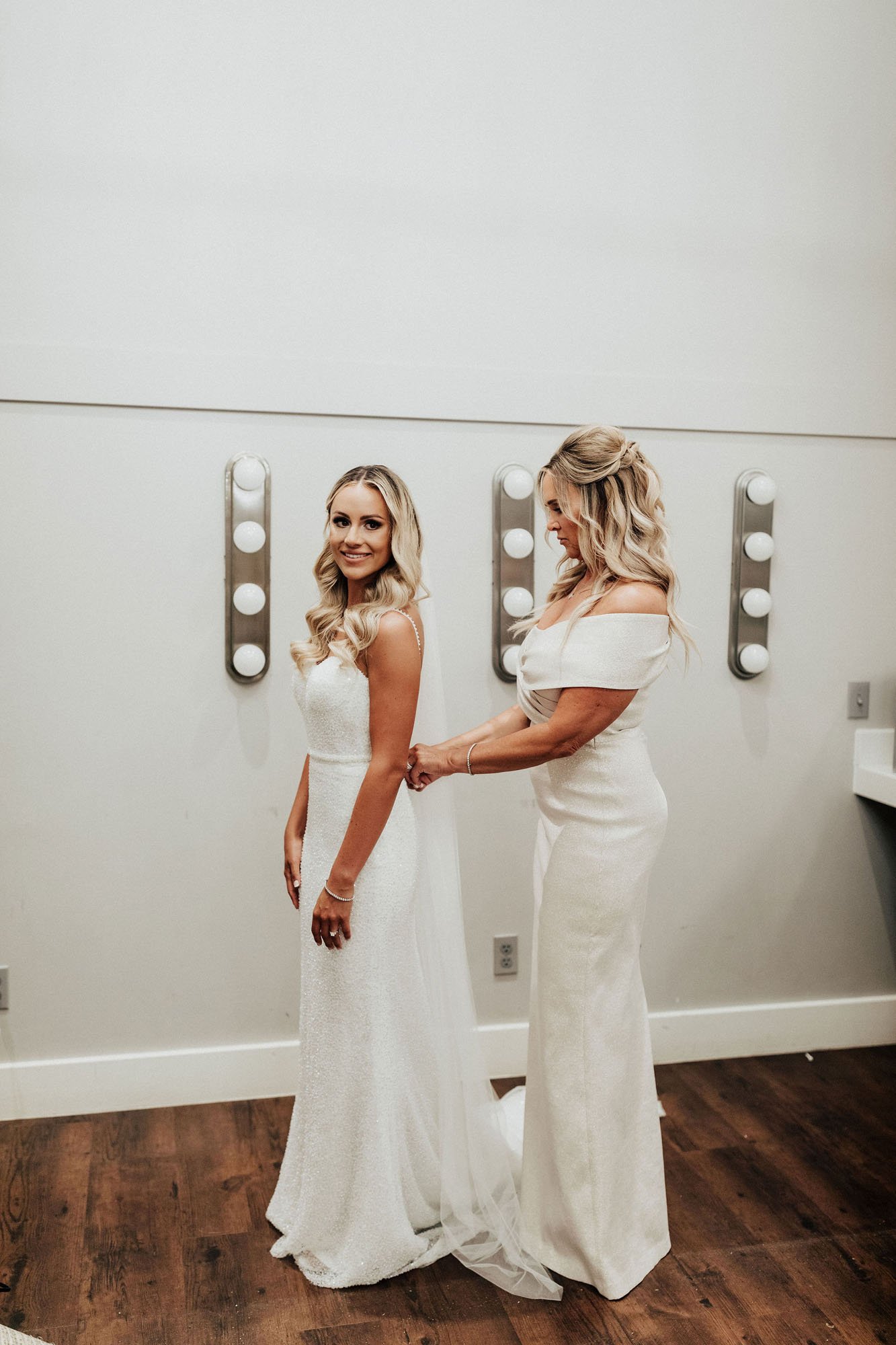  Photograph of a bride wearing Made With Love Lola while the mother of the bride helps her button her wedding dress. They both have blonde hair with soft curls, and are in a bridal suite getting ready for the wedding day at a vineyard near Seattle. 